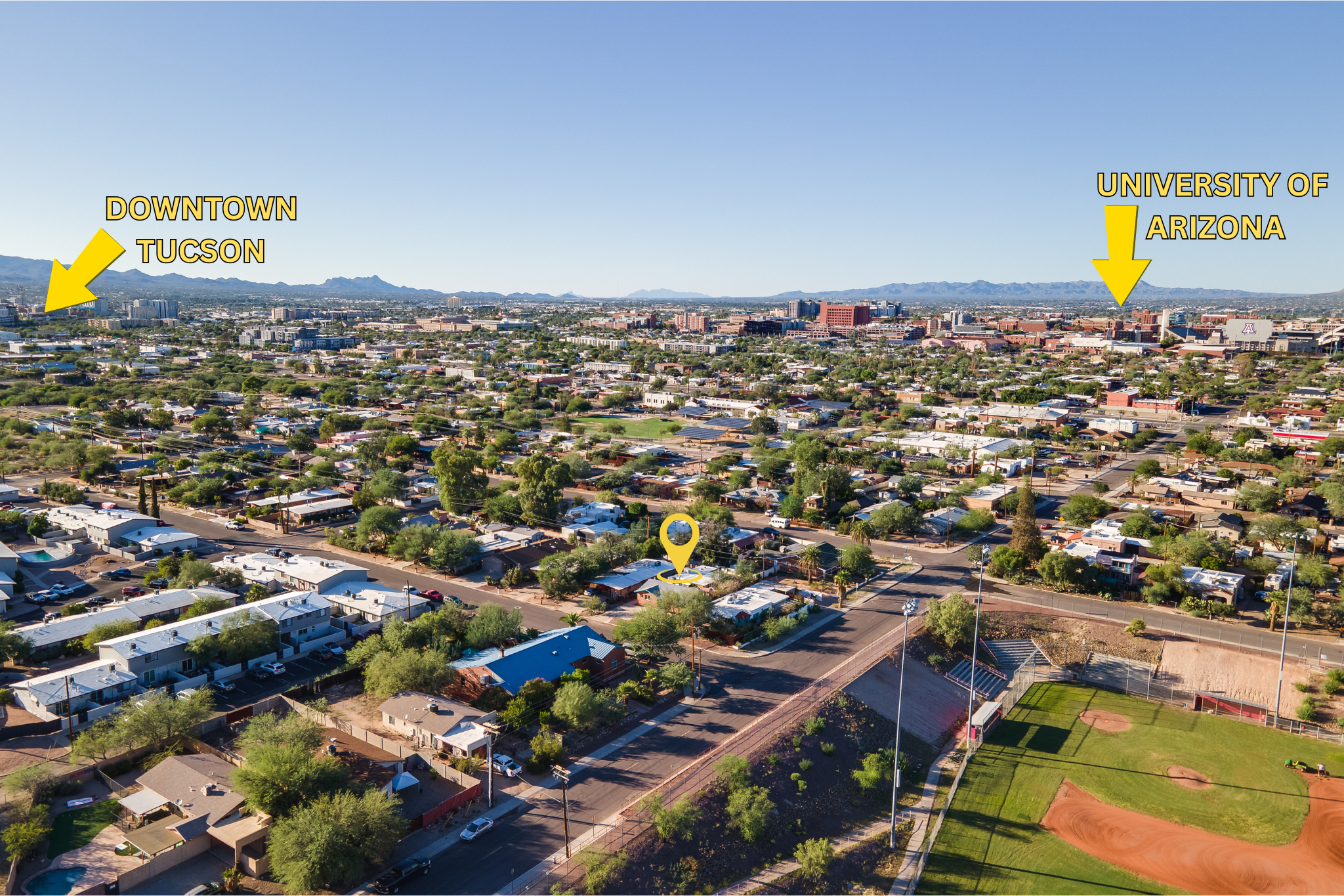 Aerial view of Tucson, Arizona, with labels pointing to downtown Tucson and the University of Arizona. Residential neighborhood in the foreground with a baseball field and sports stadium. Mountains in the background under a clear blue sky.