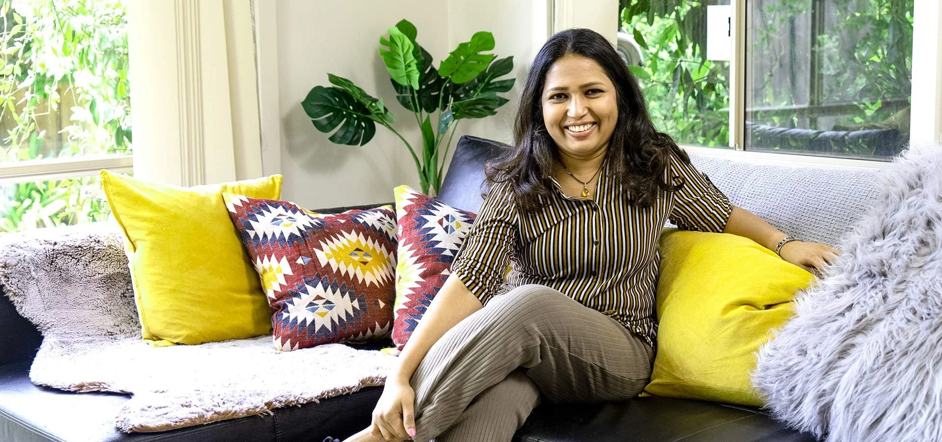 A woman sitting on a sofa surrounded by colorful pillows, with a large green plant and windows with a garden view in the background.