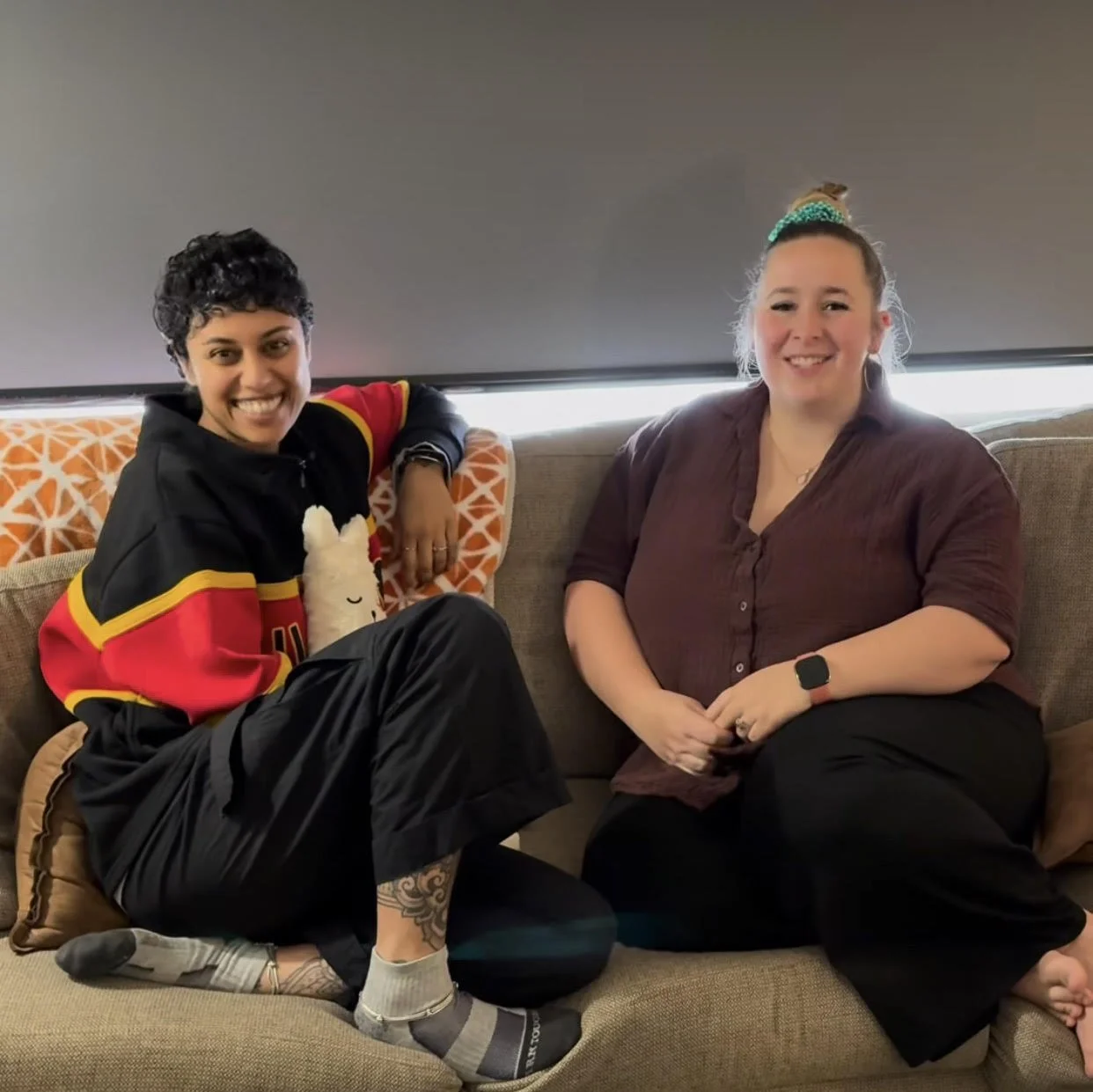 Two women sitting on a beige couch, smiling at the camera. The woman on the left has short dark hair, is wearing a black and red hoodie with black pants, and has a tattoo on her lower leg. The woman on the right has light brown hair tied up with a green scrunchie, wearing a maroon shirt and black pants, and is wearing a black smartwatch.
