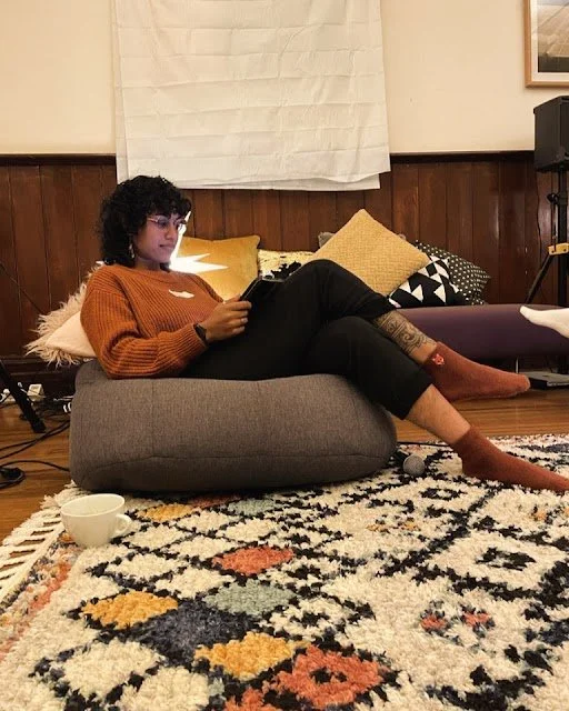 A person with curly hair and glasses sitting on a floor chair, reading a book, in a cozy room with wooden walls and various pillows, with a patterned rug and a small white bowl nearby.
