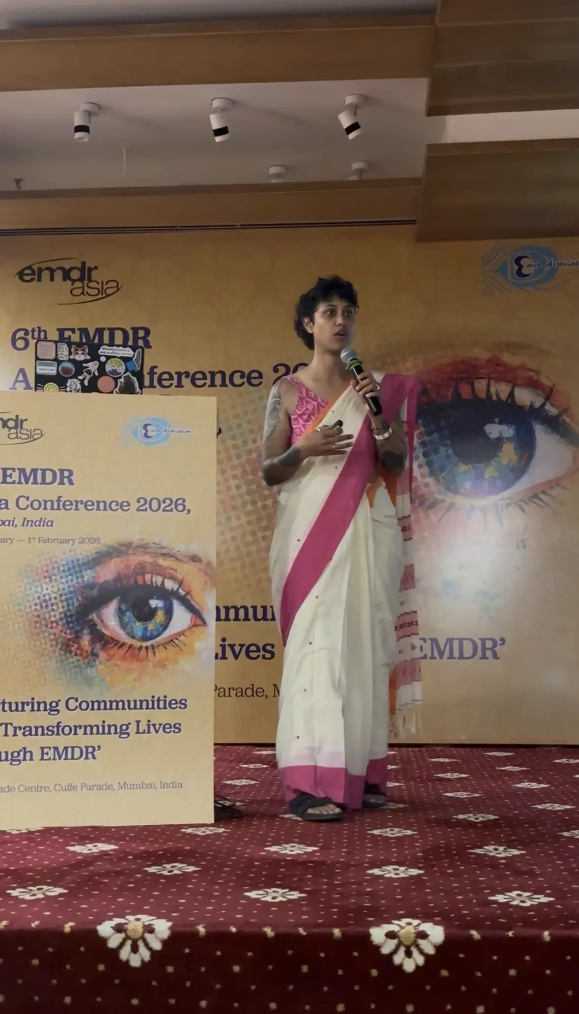 A woman dressed in a traditional white saree with pink accents is speaking into a microphone on stage at a conference. Behind her, there is a large yellow backdrop with a colorful eye illustration and text about the EMDR Asia Conference 2026 in Mumbai, India.