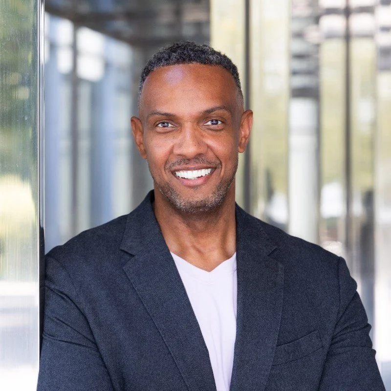 Greg Boone smiling with short curly hair, wearing a dark blazer over a white shirt, standing outdoors near glass windows.