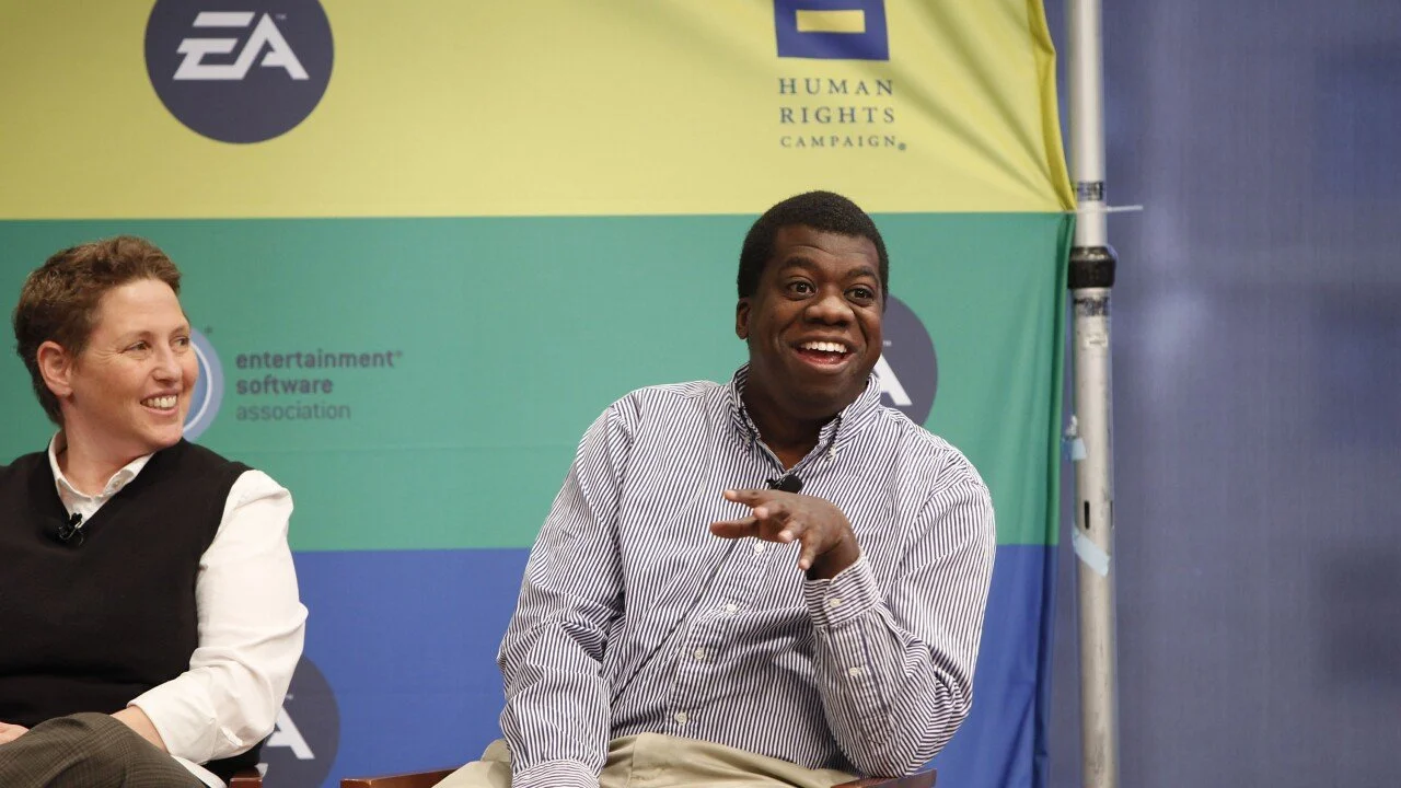 Gordon Bellamy sitting in front of a colorful backdrop with Electronic Arts (EA) and Human Rights Campaign (HRC) logos, one smiling and talking, and the other smiling and listening.