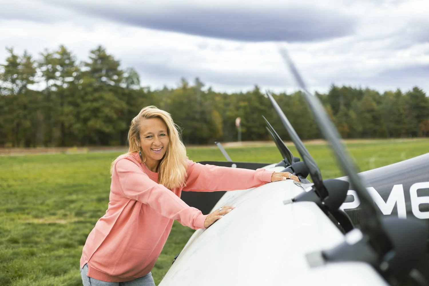Heather Chirtea in pink sweater leaning on a small aircraft outdoors on a grassy field with trees in the background.