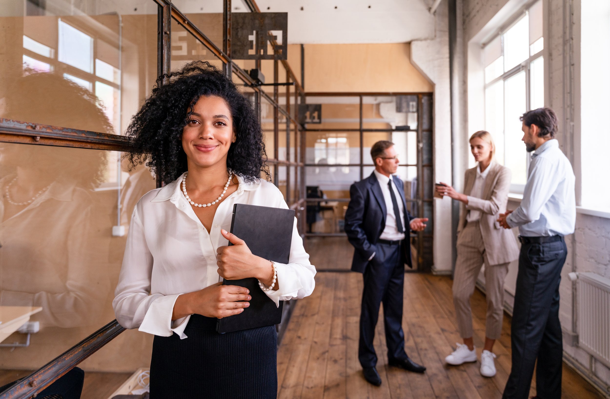 A confident woman smiling and holding a folder in a modern office space, with three colleagues engaged in a discussion in the background.