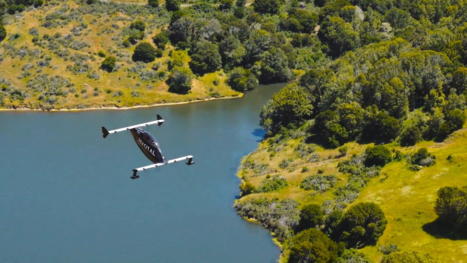 An aerial view of Heather Chirtea in the blackfly flying car over a lake surrounded by green hills and trees.
