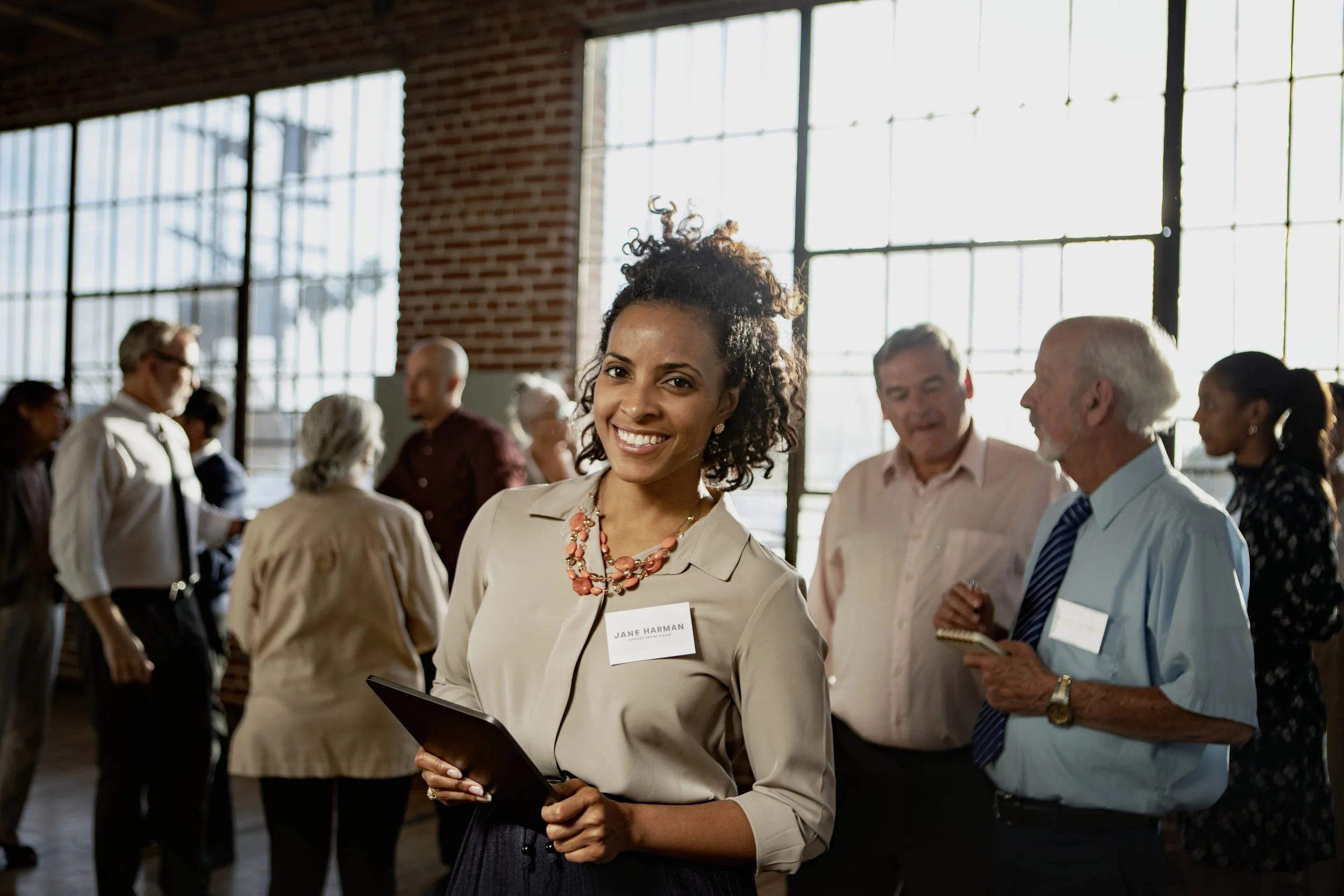 A woman smiling at a professional networking event, holding a tablet, with a name badge that reads Jane Harman, wearing a beige blouse and coral necklace, surrounded by diverse attendees in a bright, industrial-style venue.