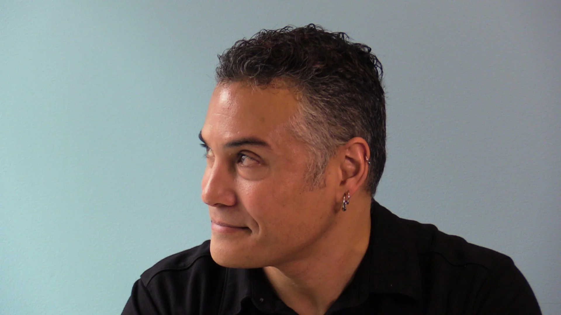 Omar Abdelwahed with short, curly, dark hair with gray streaks, wearing earrings, looking to his left. He is dressed in a black shirt and sitting against a plain, light-colored background.