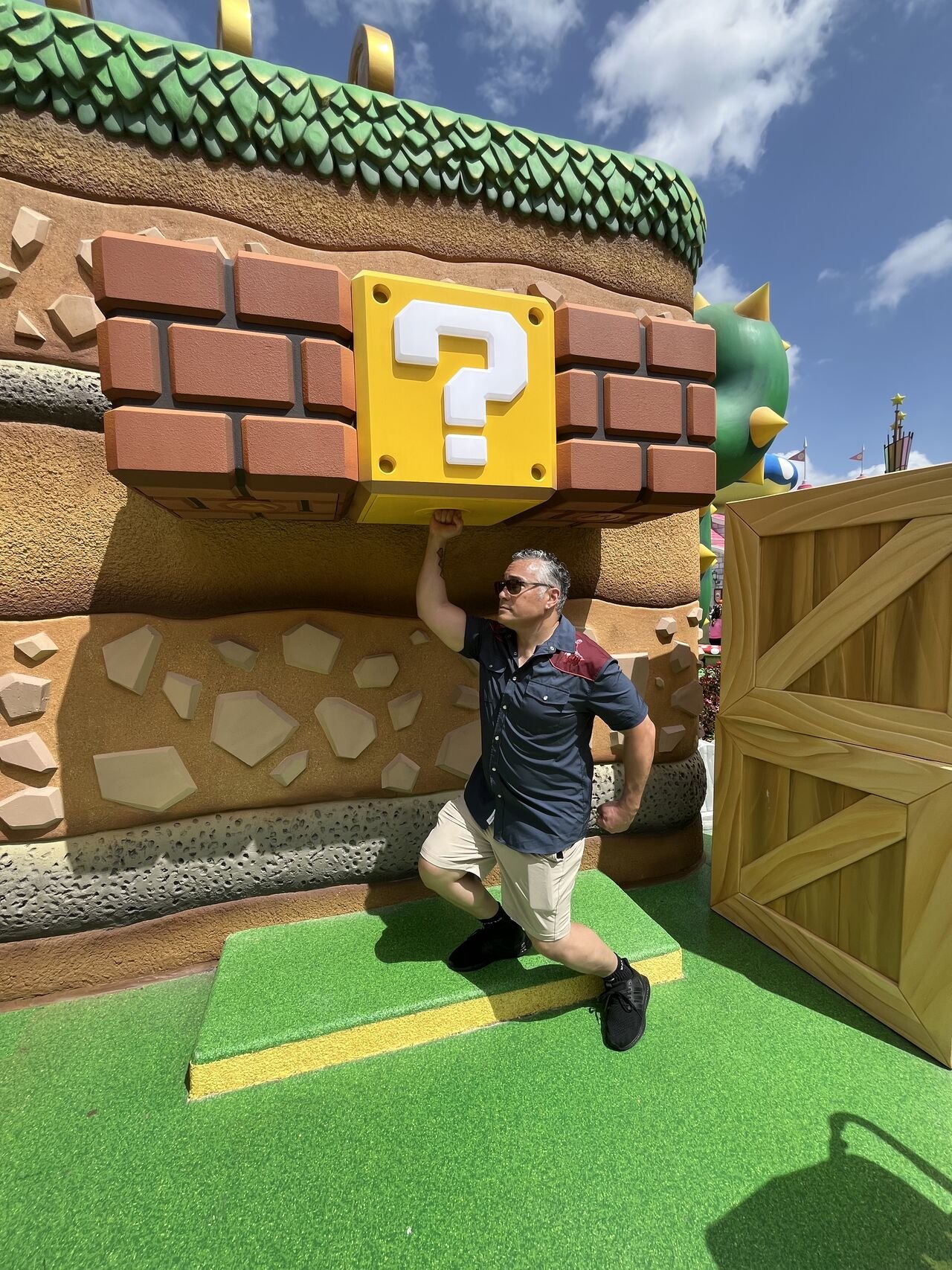 Omar Abdelwahed posing with one arm raised, underneath a large yellow block with a white question mark, part of a colorful, cartoonish structure resembling a mushroom house from Super Mario, on a bright, sunny day.