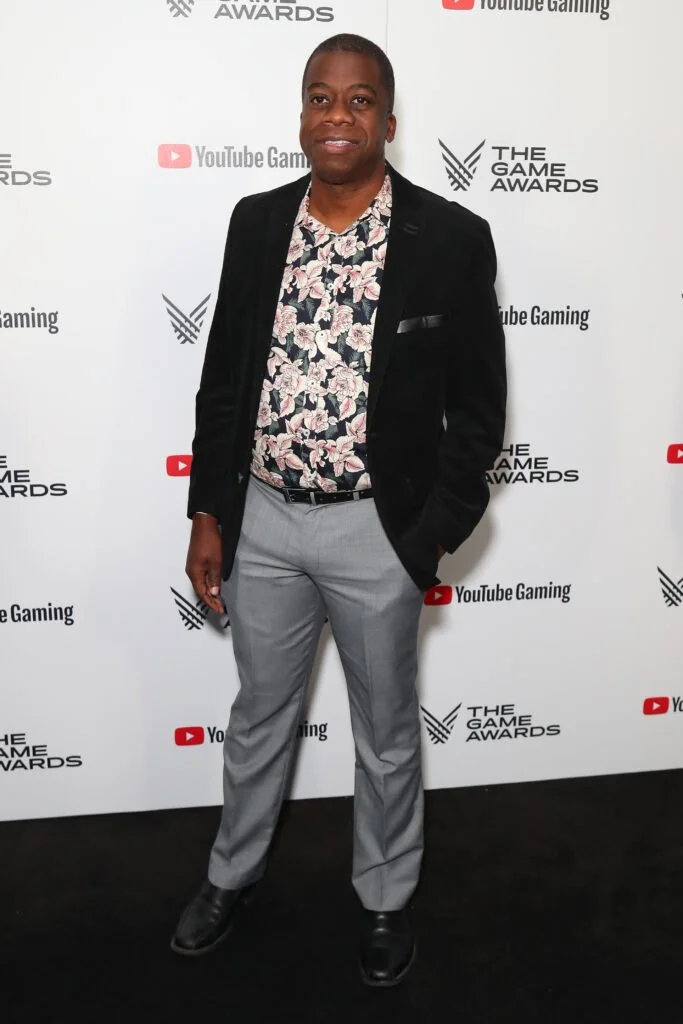 Gordon Bellamy standing in front of a backdrop with logos for YouTube Gaming and The Game Awards, wearing a floral shirt, gray pants, black shoes, and a black blazer.