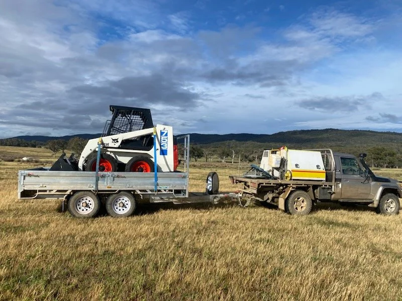 Trailer carrying a skid-steer loader, attached to a ute in a grassy field.