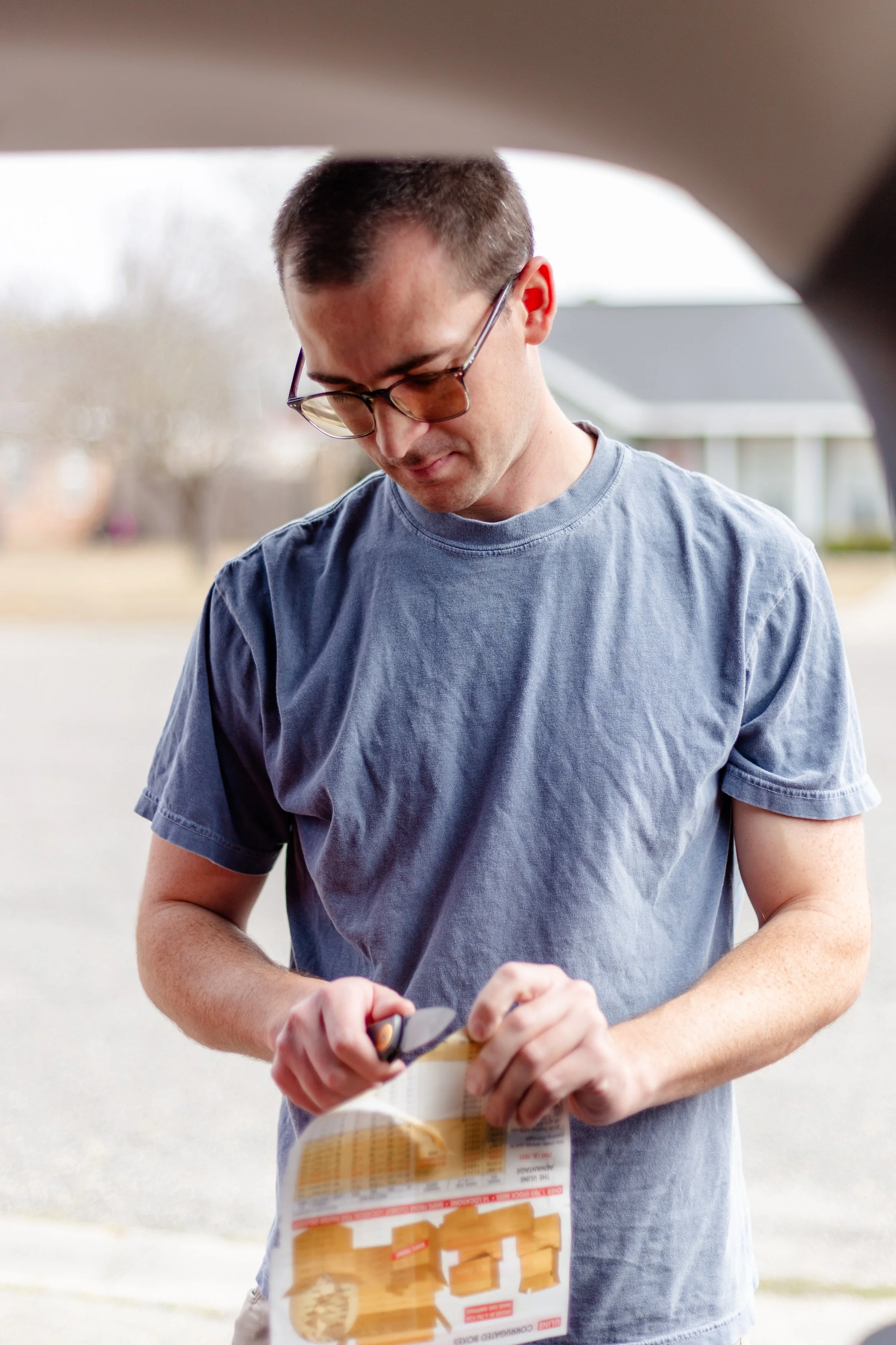 A man with short brown hair and glasses, wearing a grey T-shirt, cutting a piece of catalog paper with a knife outside during daytime.