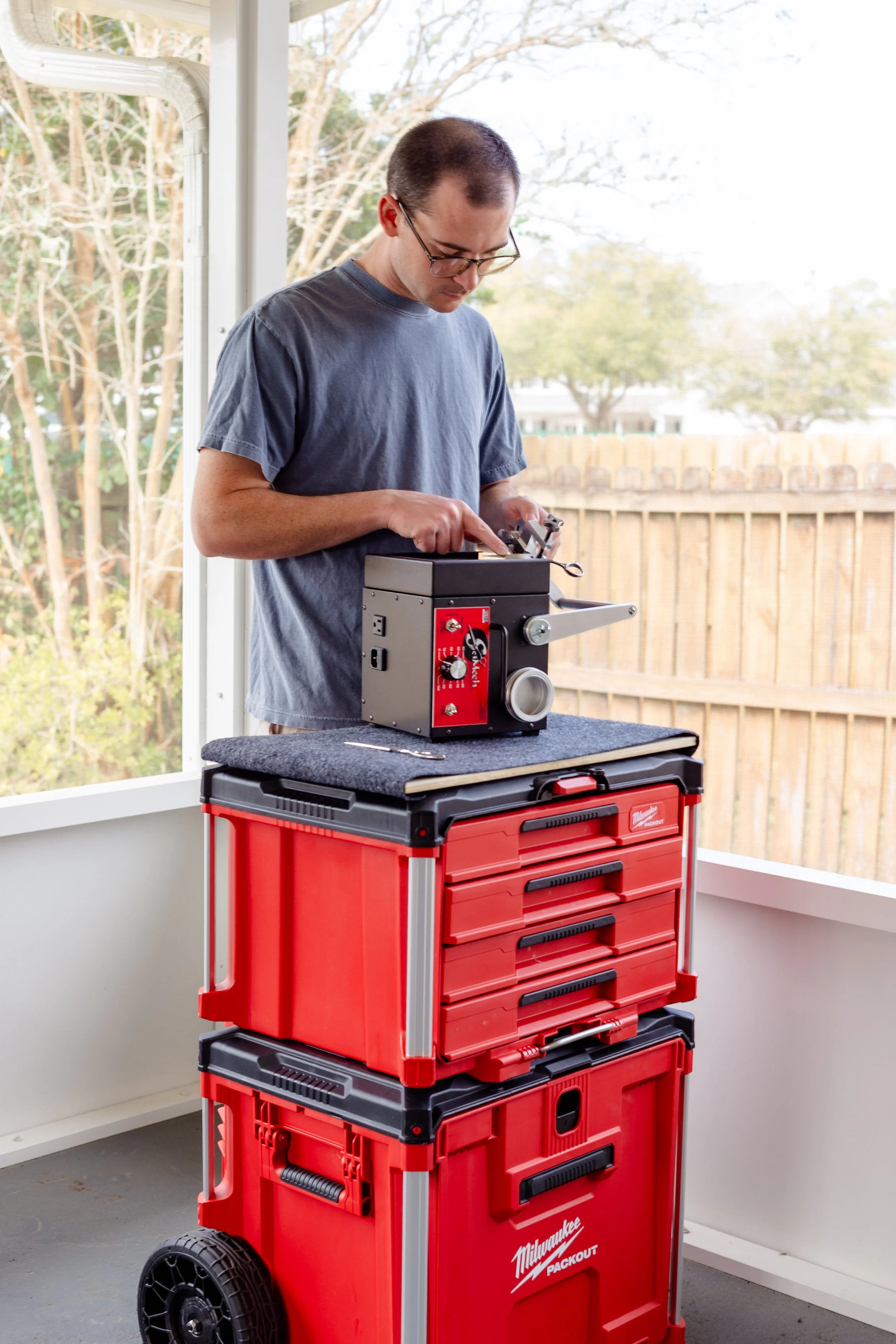 A man wearing glasses and a blue t-shirt working with a flat-hone grinder to sharpen salon shears on a tool box with a table top near a window, with a fenced backyard visible outside.