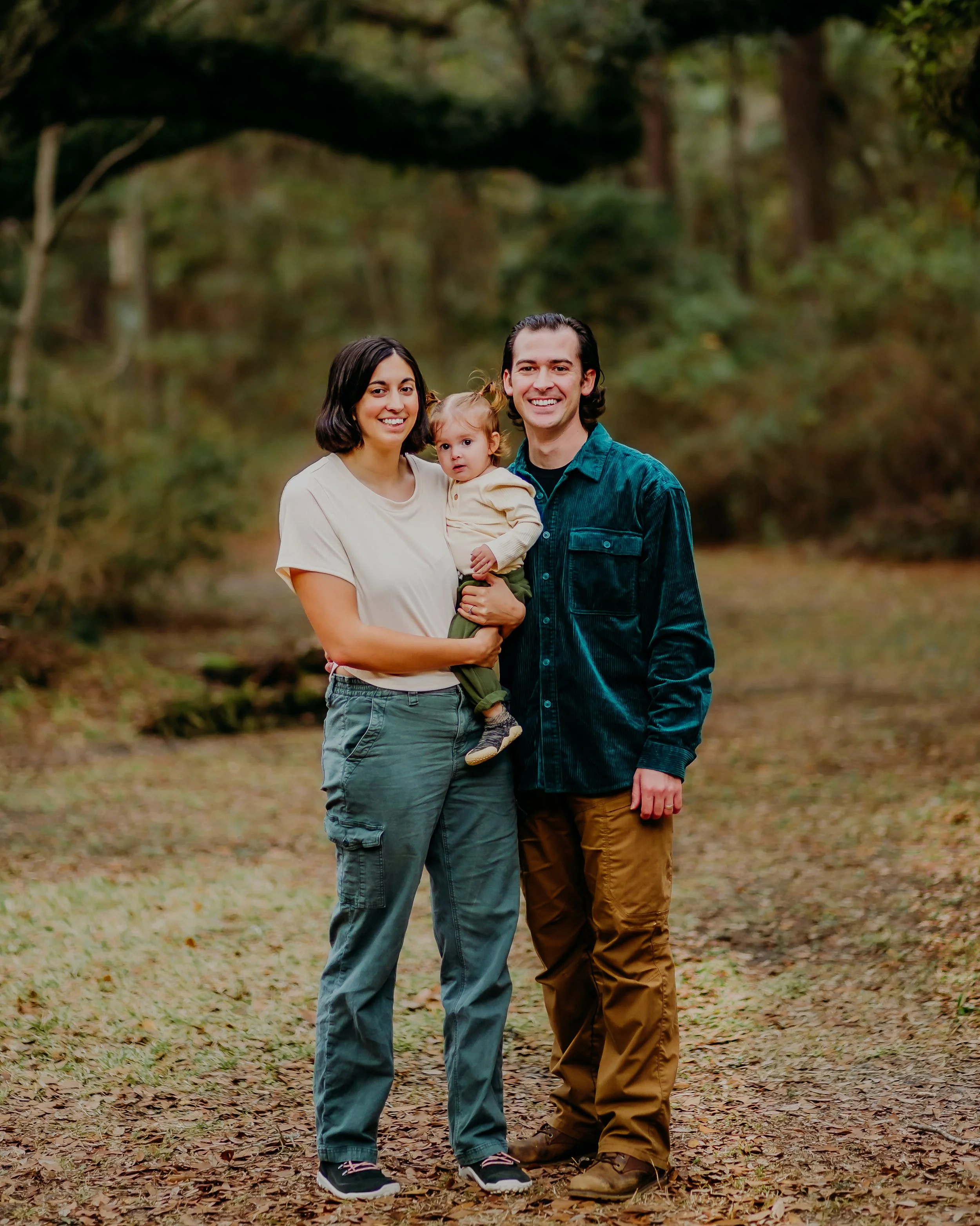 Three people standing outdoors in a wooded area, a woman holding a young girl and a man standing beside them, all smiling at the camera.