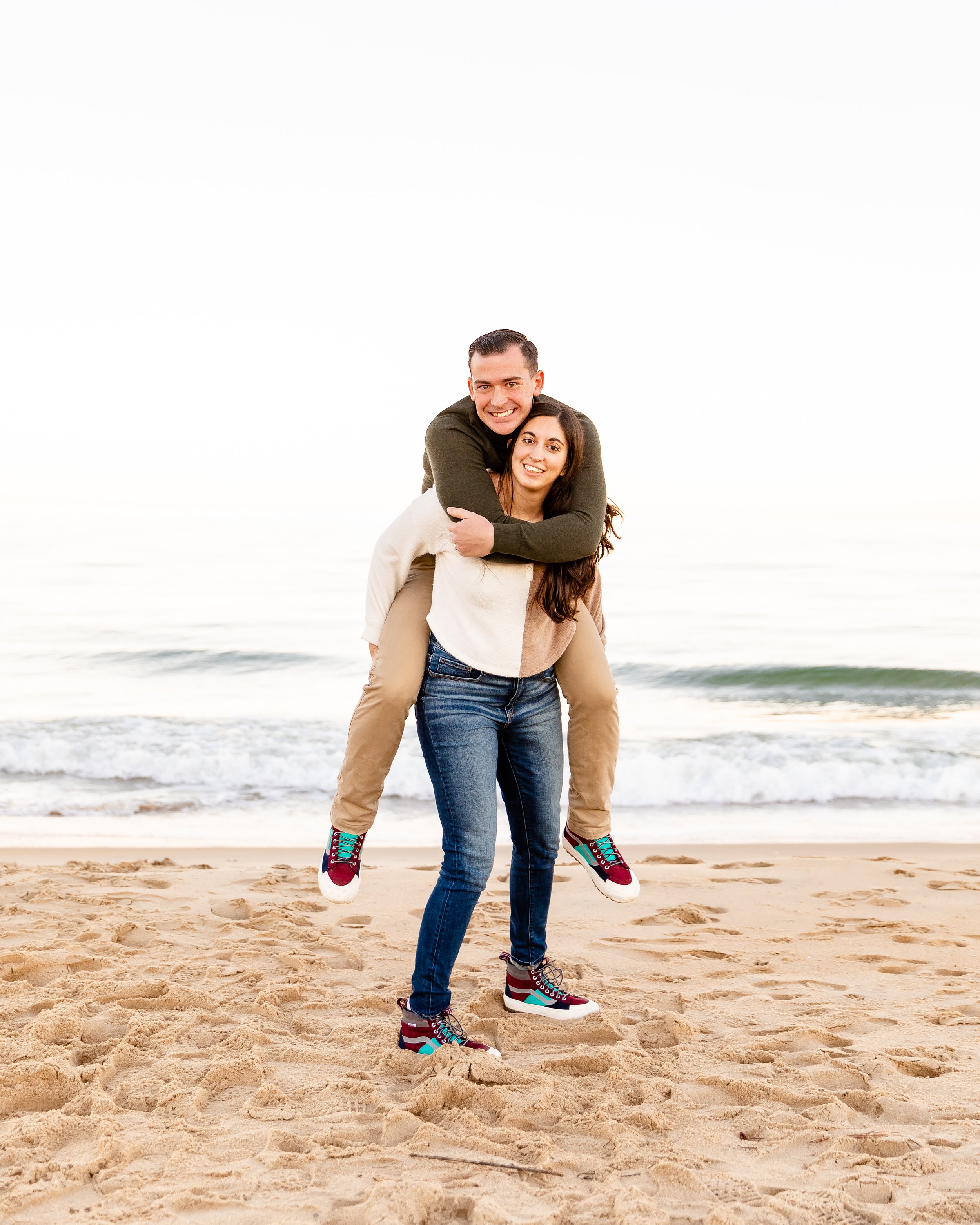 A woman carrying a man on her back at the beach during sunset, with waves in the background and sand in the foreground.
