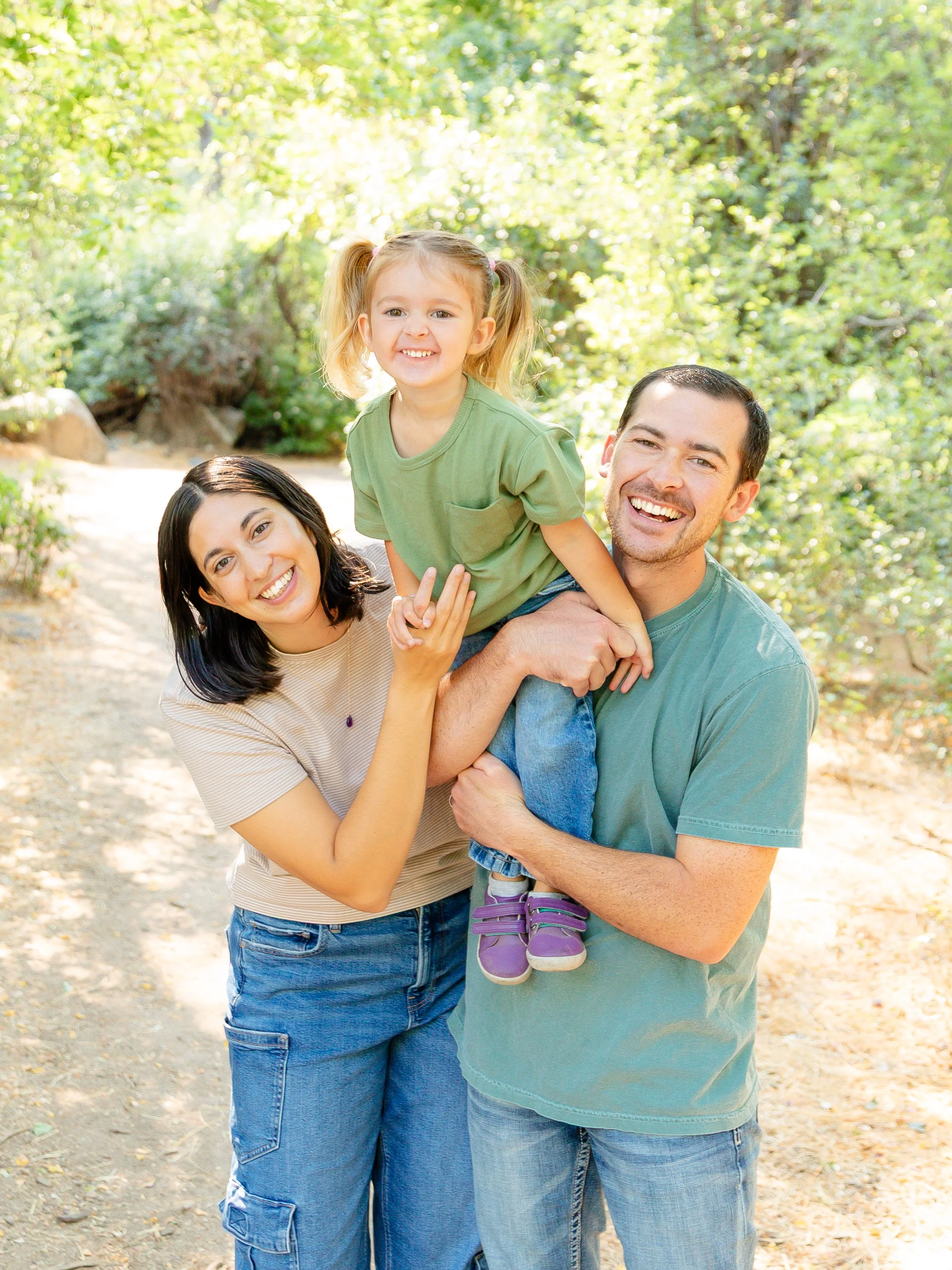 Family of three enjoying a walk outdoors, with the father holding the daughter, mother smiling, surrounded by trees and sunlight.