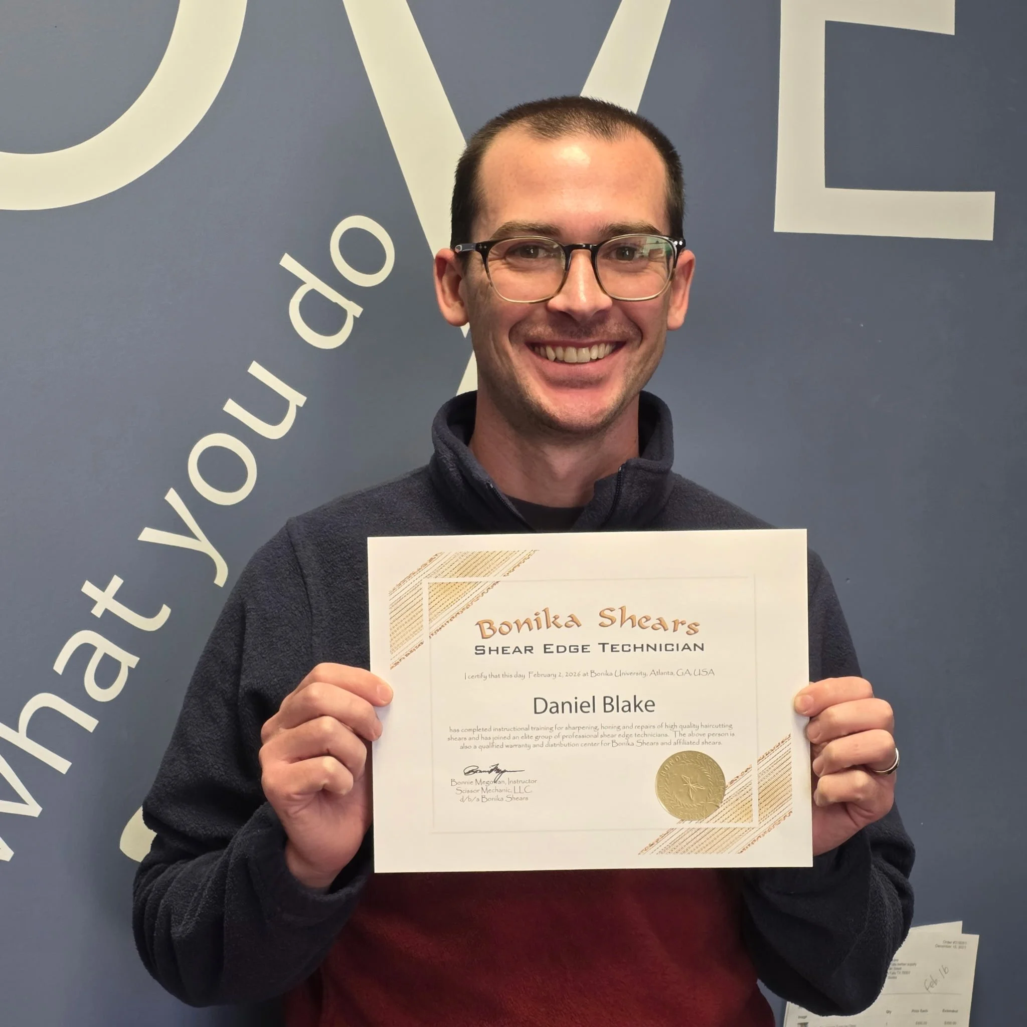 A smiling man with glasses holding a certificate, standing in front of a blue wall with partially visible large white text.
