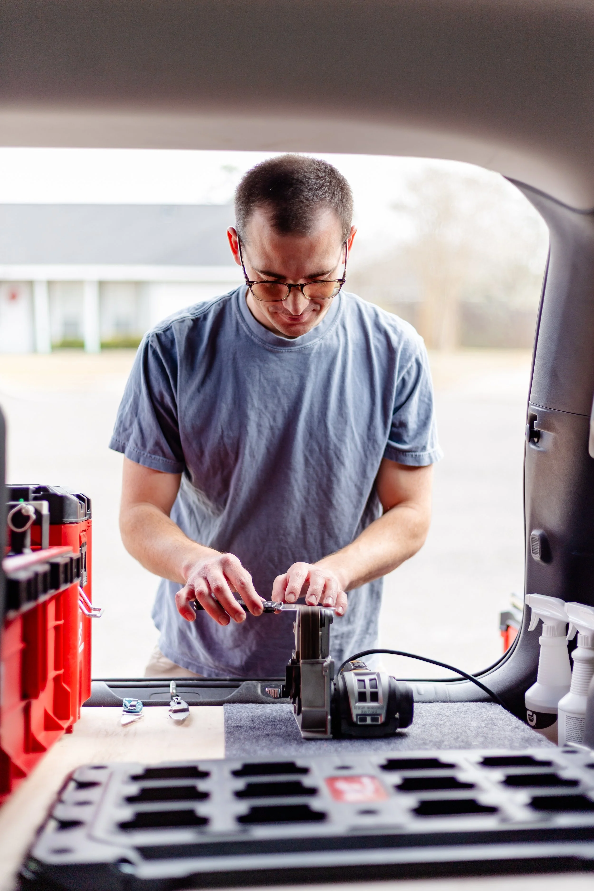 A man with glasses sharpening a knife in the back of a vehicle, with tools and supplies on a work surface.