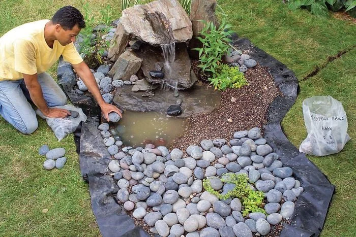 A person working on a backyard landscaping project, arranging gray stones around a small pond with a waterfall feature, surrounded by a black landscape fabric, plants, and grass.