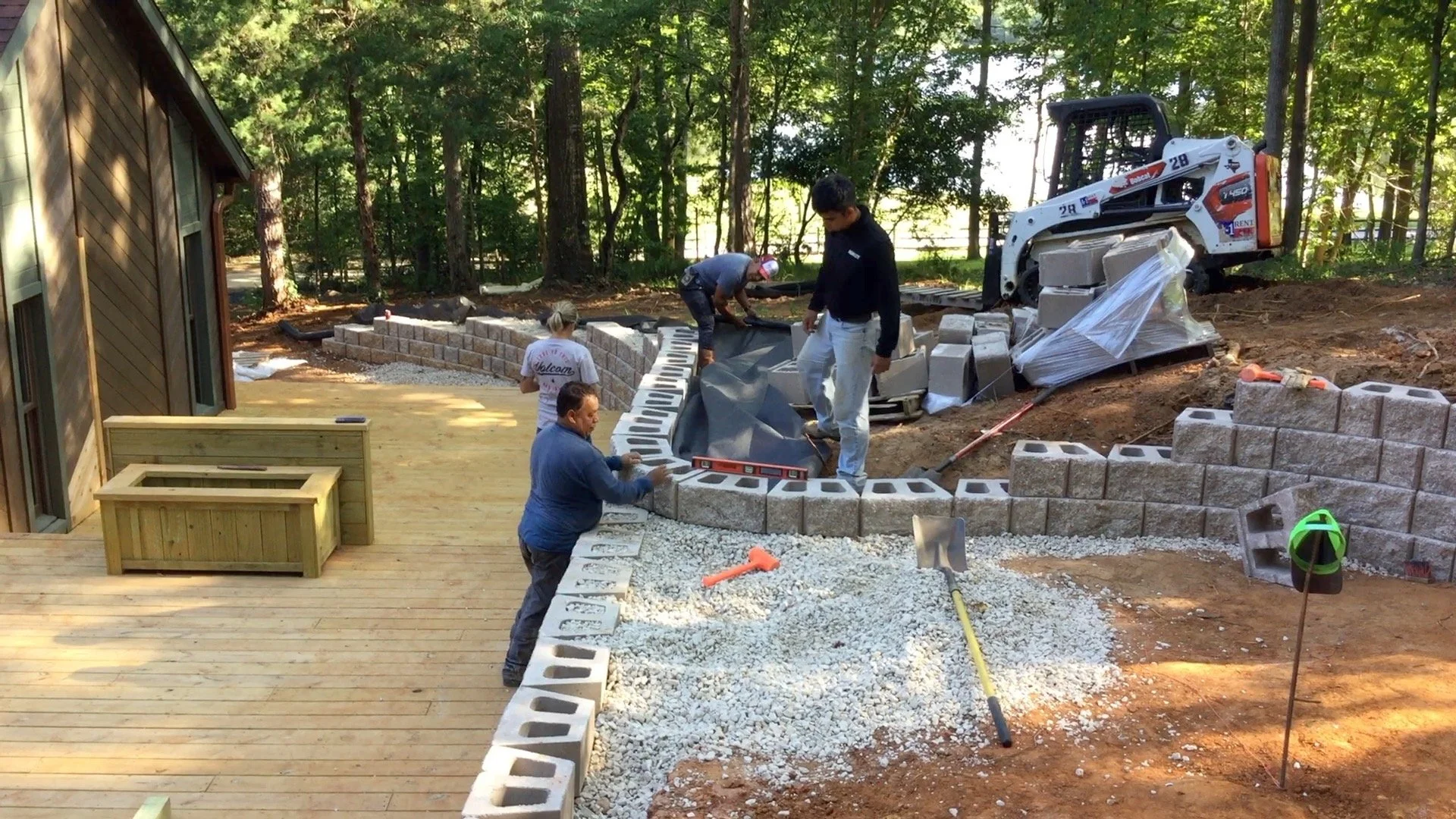 People constructing a brick wall for a landscape project outdoors near a house, with trees in the background. Some are placing bricks, others are handling materials, with construction tools visible.