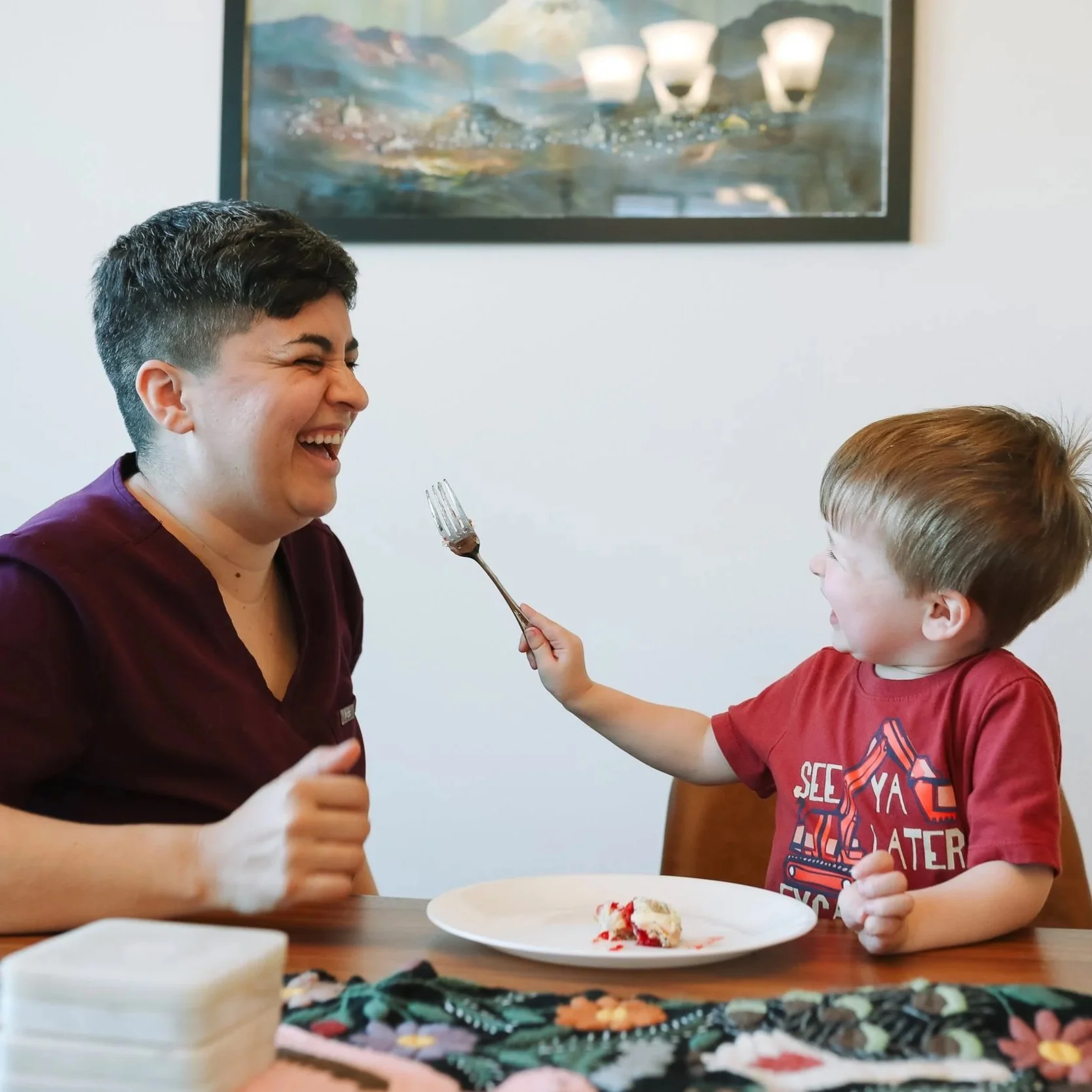 A young boy wearing a red t-shirt feeding a woman with a fork, both smiling and laughing while at a table with a plate of dessert in front of them, in a room with a framed painting on the wall.