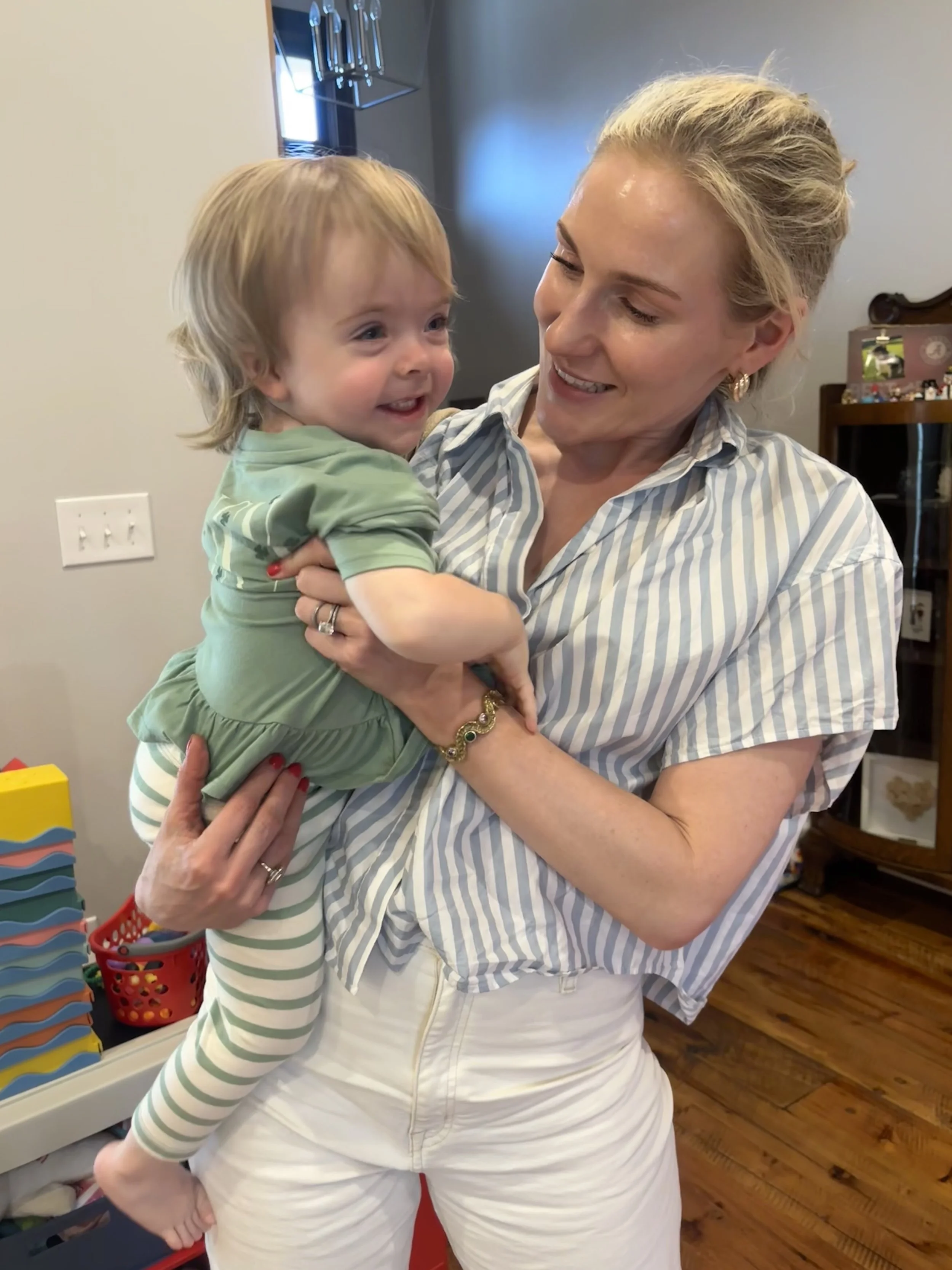 A woman holding a smiling toddler in a room with toys and a wooden floor.