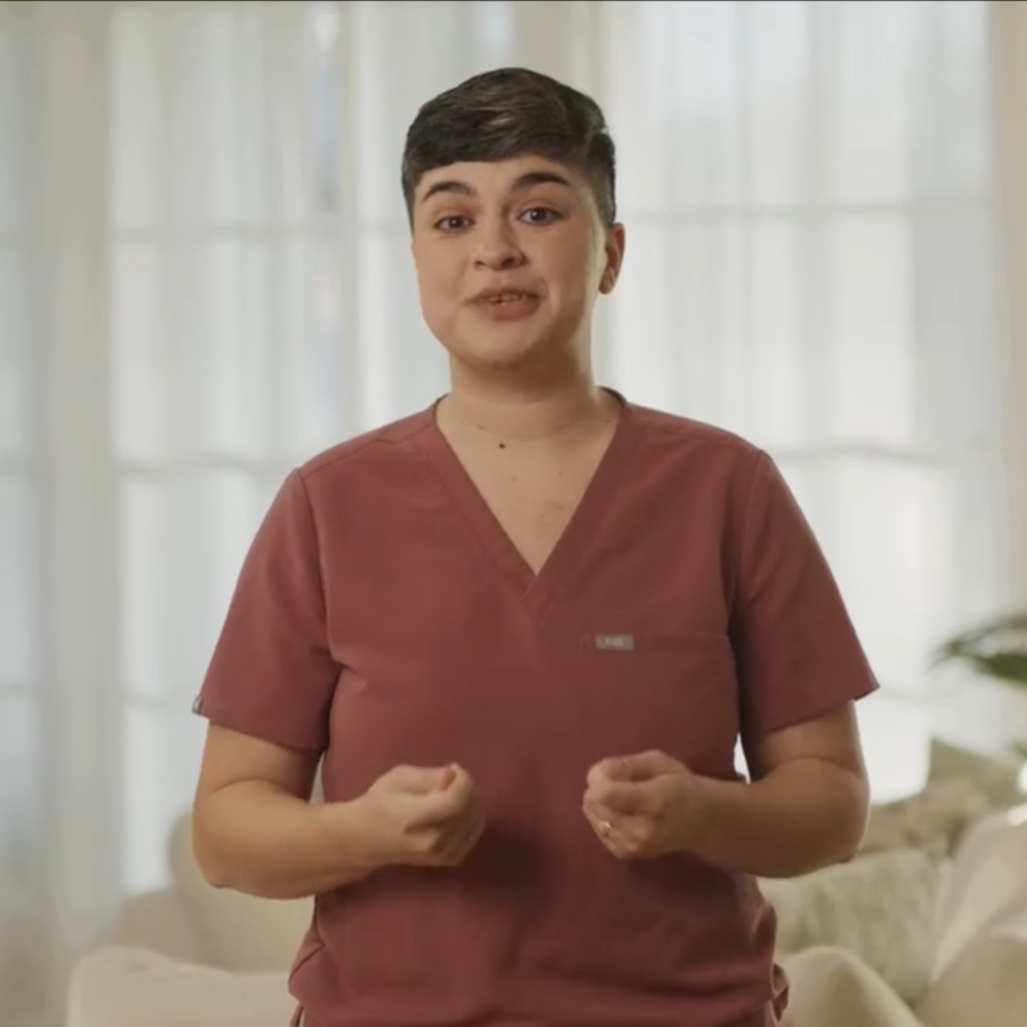 Marissa Ramos, speech therapist in a reddish-brown scrub top standing in a well-lit room with light-colored curtains in the background.