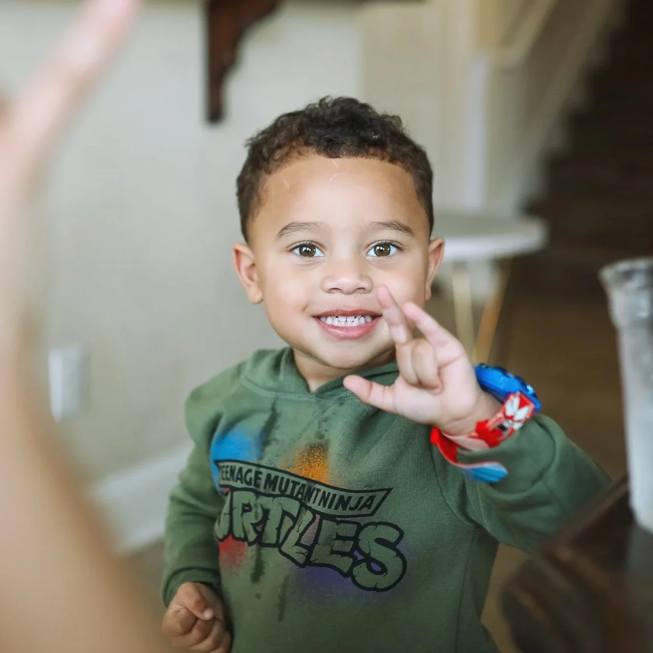 Smiling young boy with short curly hair making the "I love you" sign with his fingers, wearing a Teenage Mutant Ninja Turtles t-shirt and a red and blue wristband, indoors with stairs in the background.