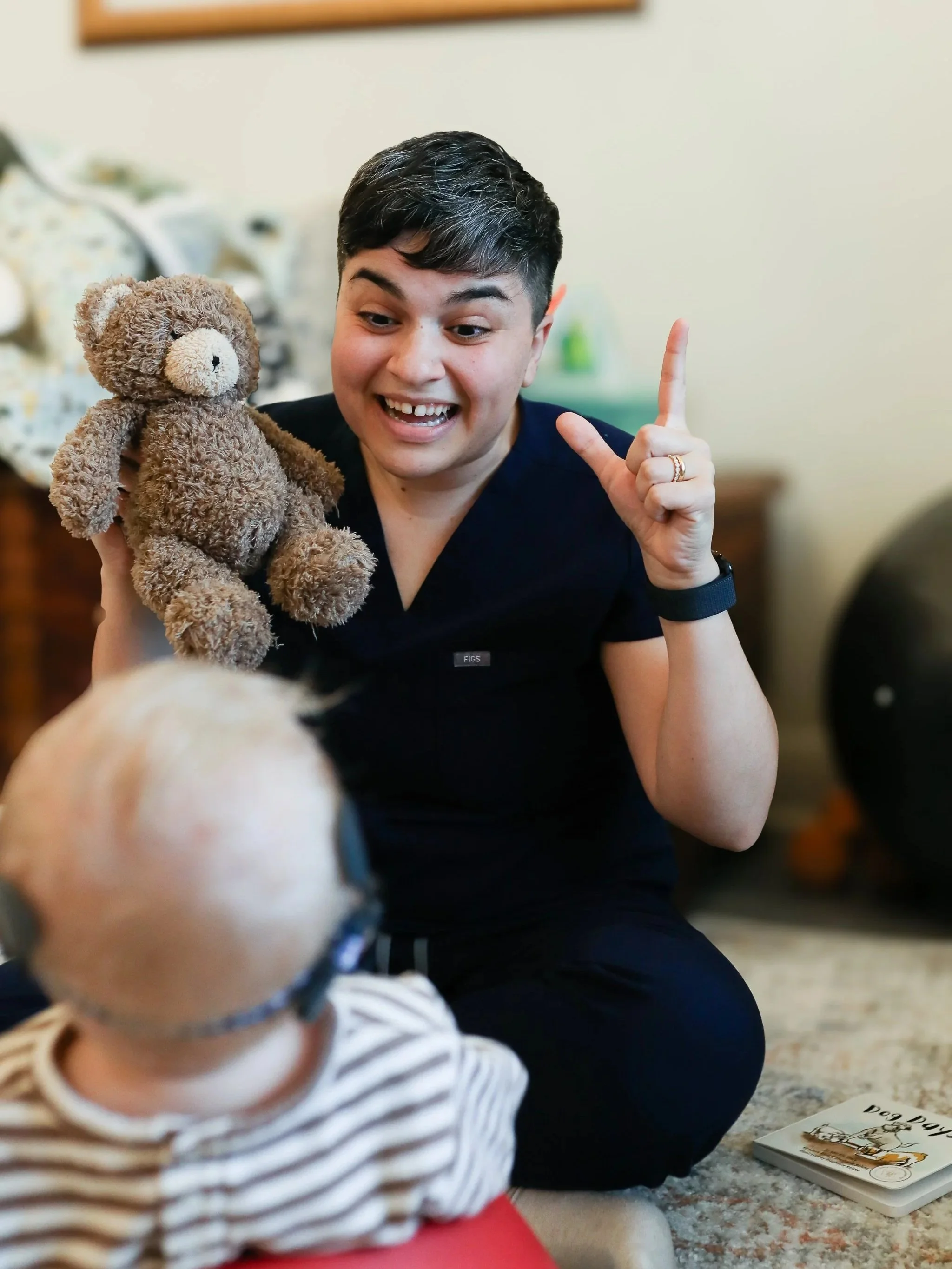 Young woman with short dark hair, smiling, holding a brown teddy bear, and making an 'L' sign with her hand. A child in striped clothing and glasses is in the foreground, with a dog book on the carpet nearby.