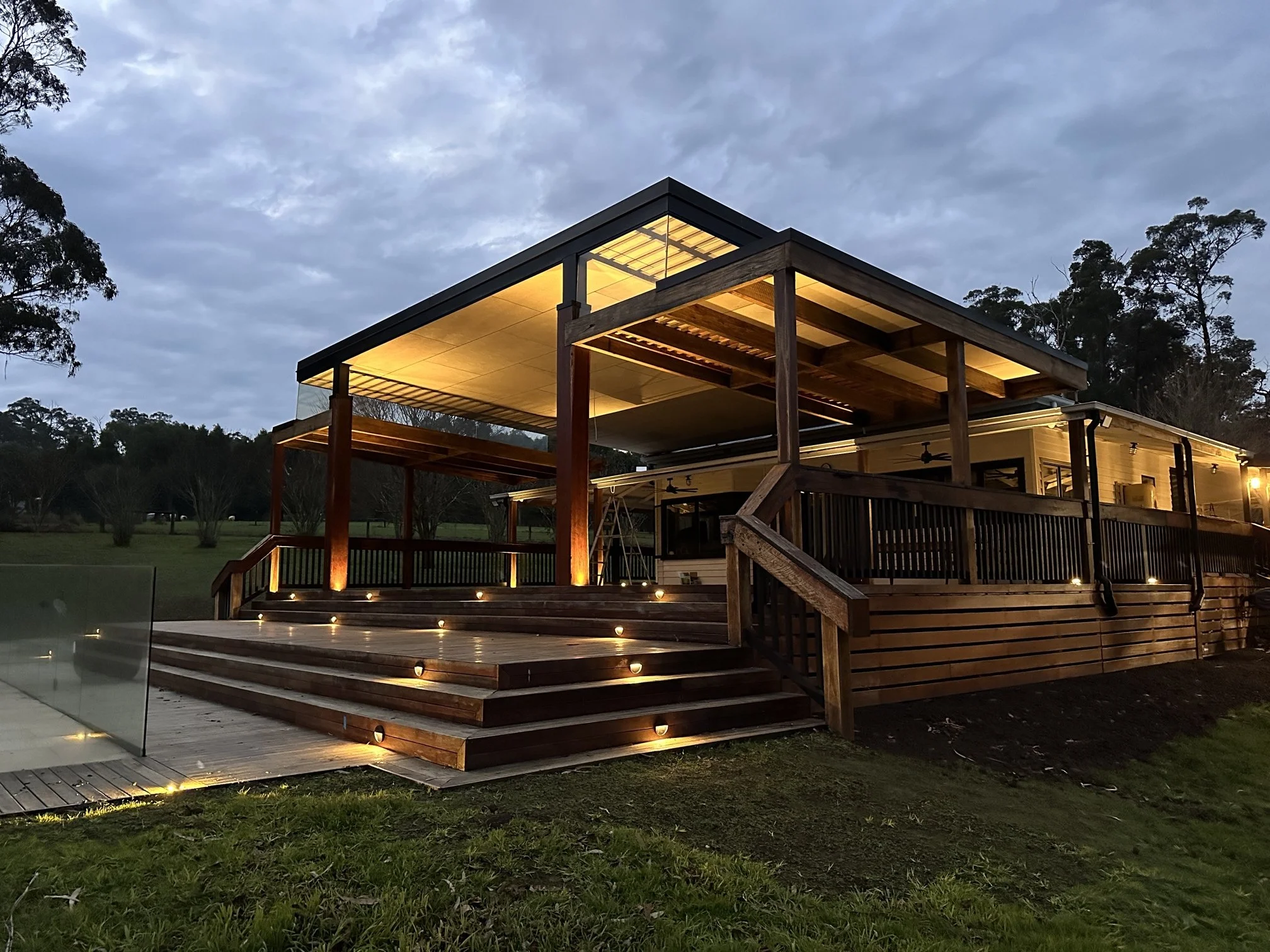 Modern two-story house with wooden exterior, illuminated with outdoor lighting, featuring a large deck and stairs, set against a cloudy sky and trees at dusk.