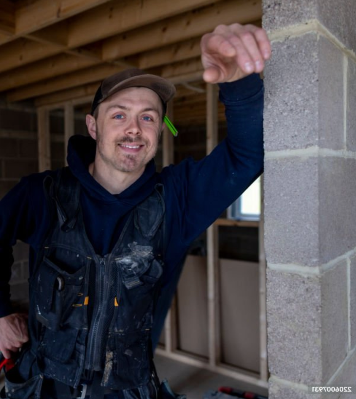 A smiling construction worker in a black vest with paint splatters, a brown cap, and a black hoodie, raising his right arm in front of a stone wall inside a building under construction.
