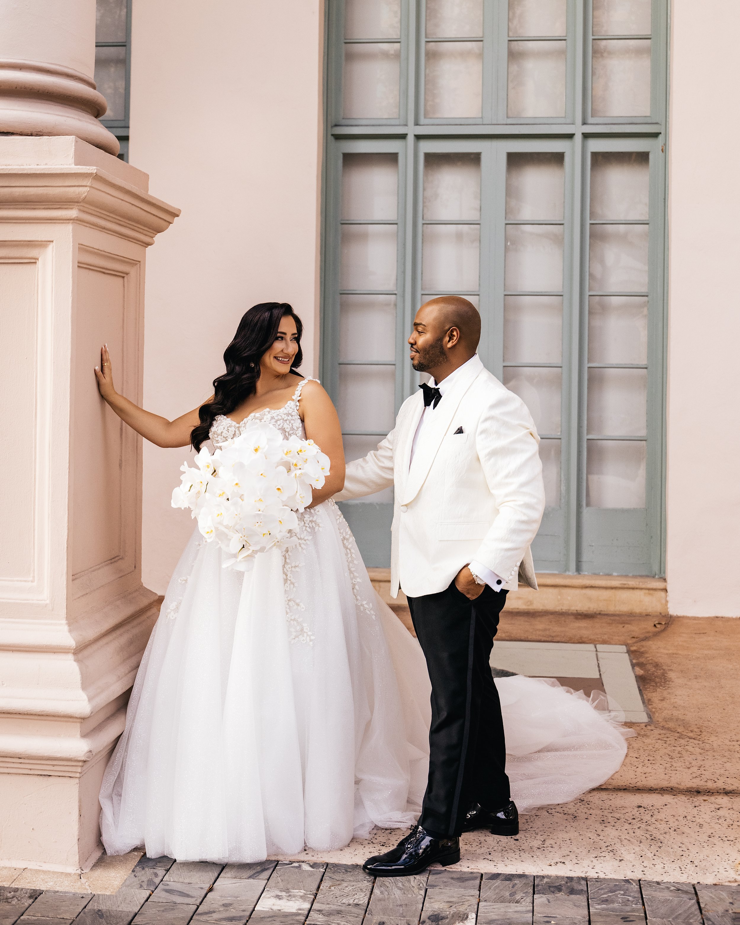 A bride and groom in wedding attire smiling at each other indoors, with the bride holding a bouquet of white flowers and standing near a pink column and large blue window.