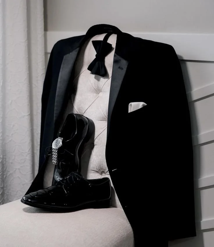 A black tuxedo jacket with satin lapels, a white dress shirt, a black bow tie, a white pocket square, a pair of shiny black dress shoes, and a black dress shoehorn placed on a beige upholstered chair.