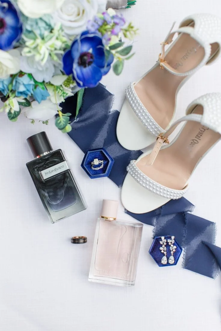White high-heeled shoes with pearl embellishments, a bouquet of white and purple flowers, perfume bottles, a jewelry box with earrings, and wedding bands arranged on a white surface.