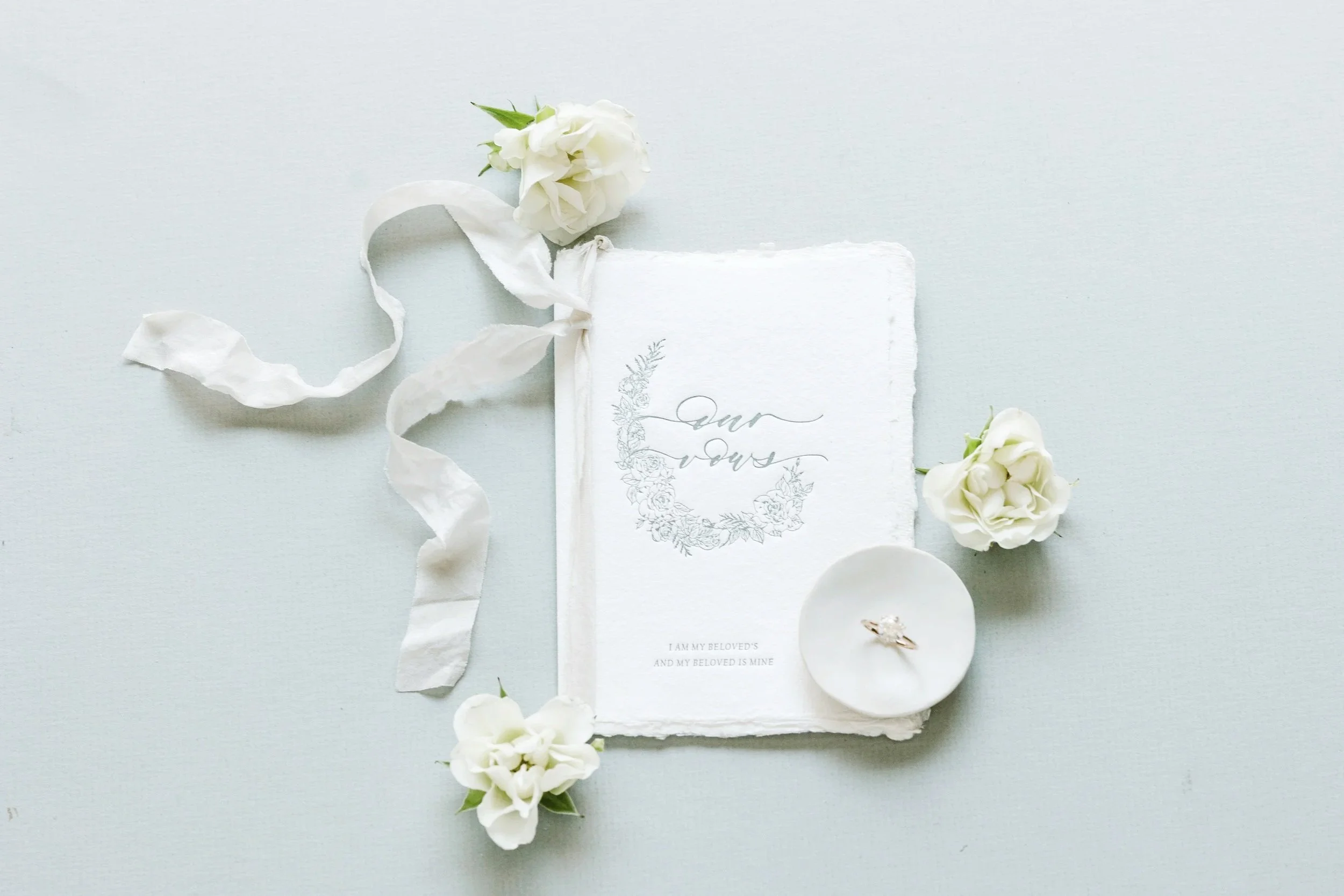 A wedding invitation with the words "Our vows" on it, surrounded by white flowers, a wedding ring on a small dish, and a white ribbon, all on a light-colored surface.