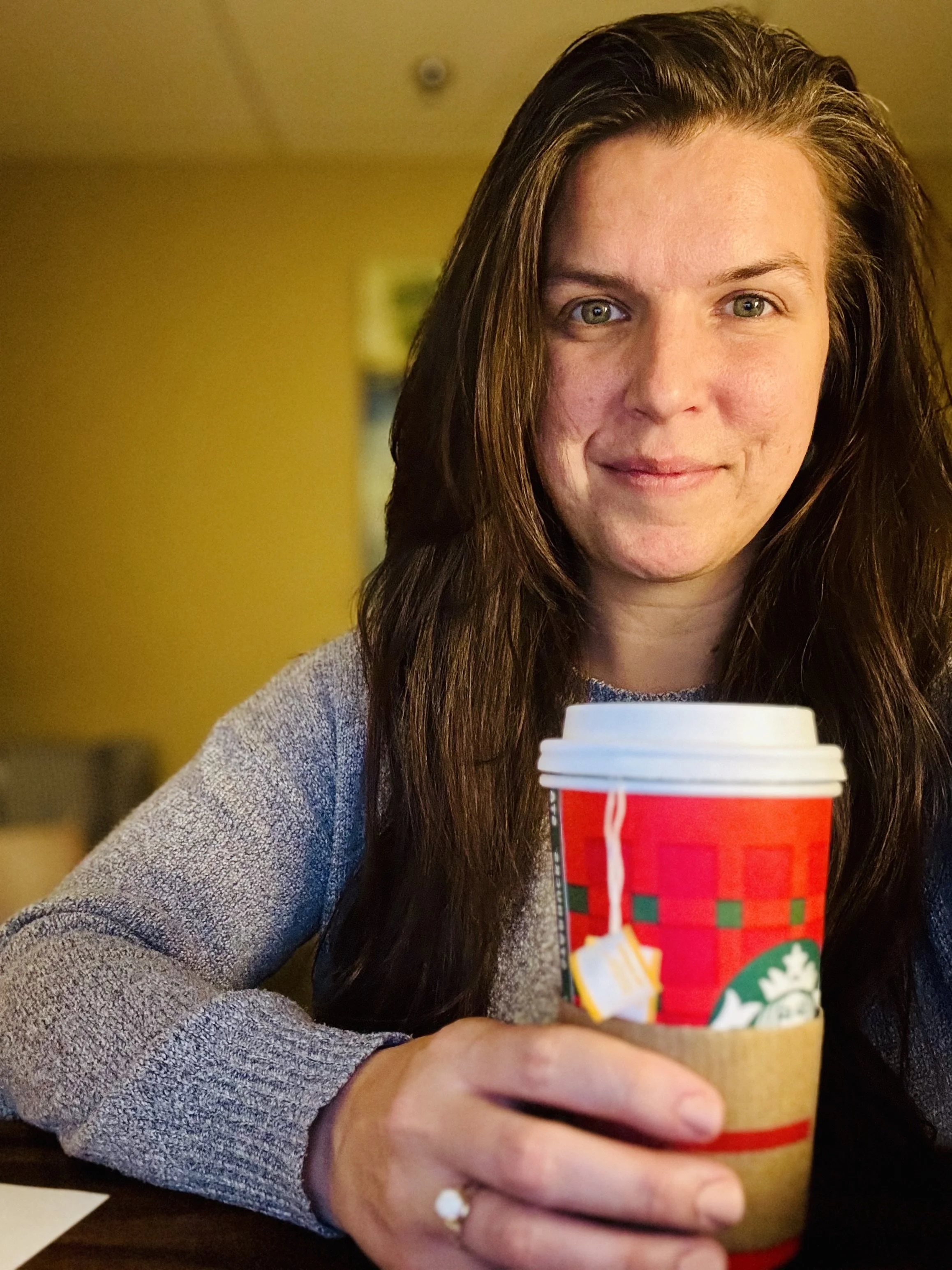 A woman with long brown hair holding a Starbucks holiday-themed coffee cup with a cardboard sleeve, smiling at the camera inside a cozy indoor setting.
