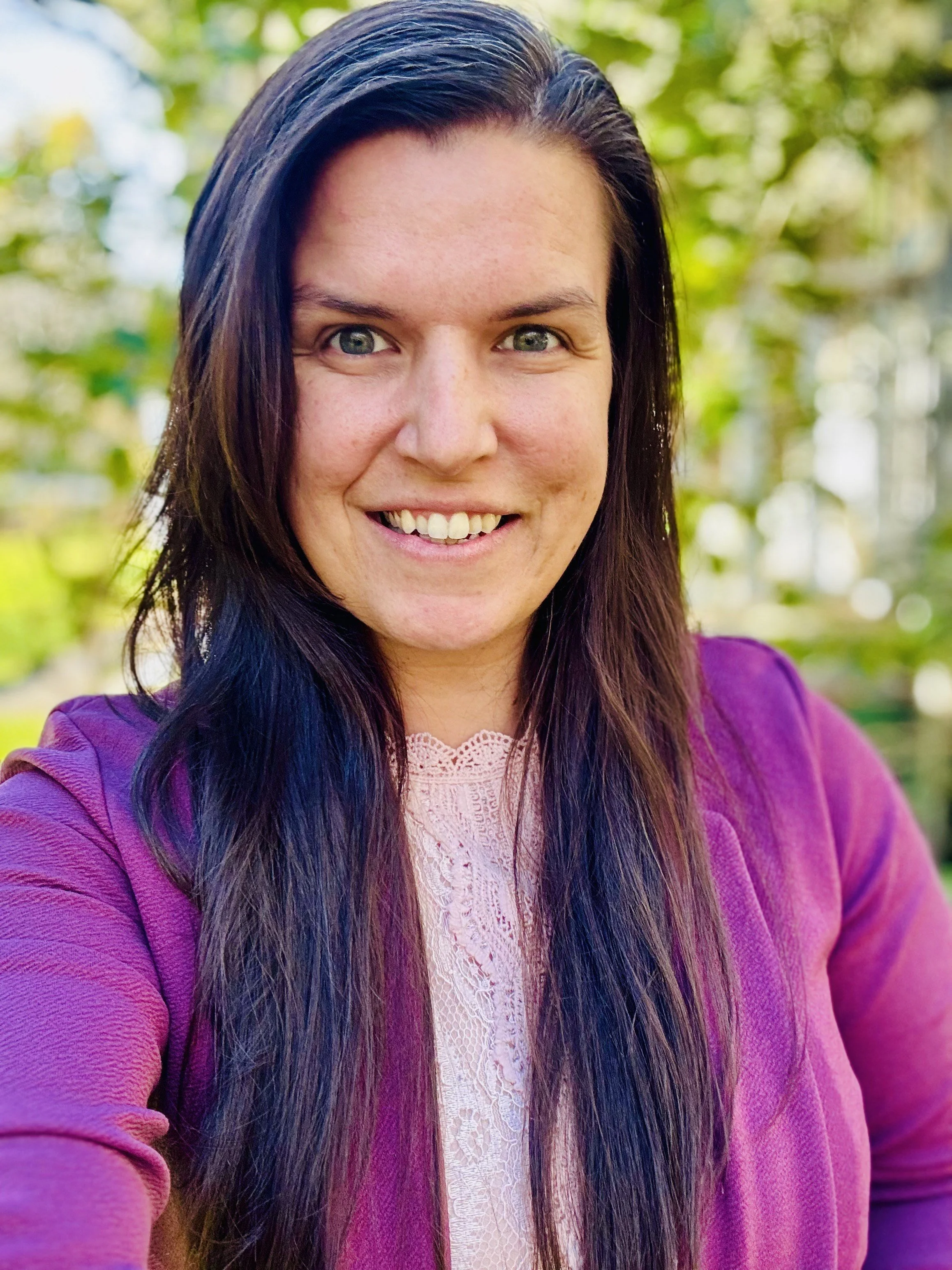 A woman with long dark hair and green eyes smiling outdoors, wearing a purple jacket and a light pink lace top.