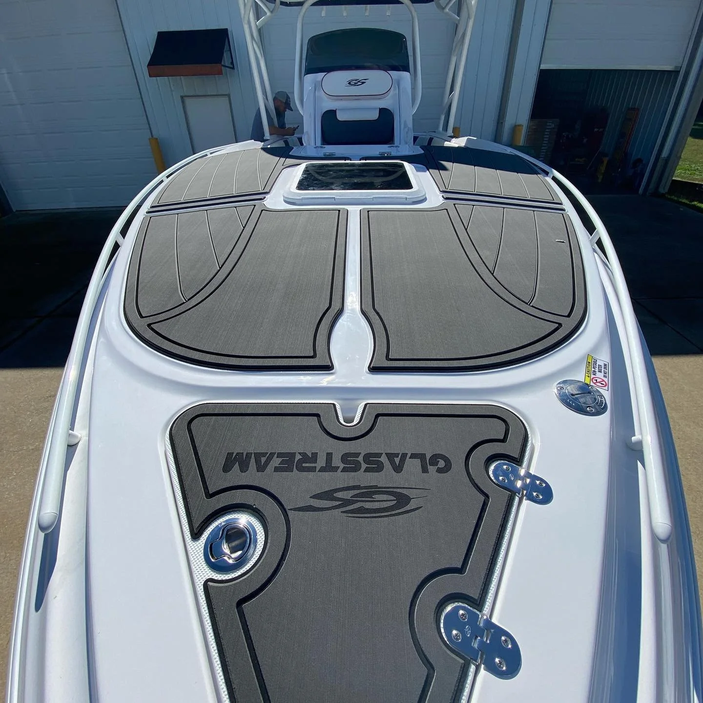 Top view of a Glastron boat with gray sun pads and a hatch in the middle, parked outside near a building with white siding.