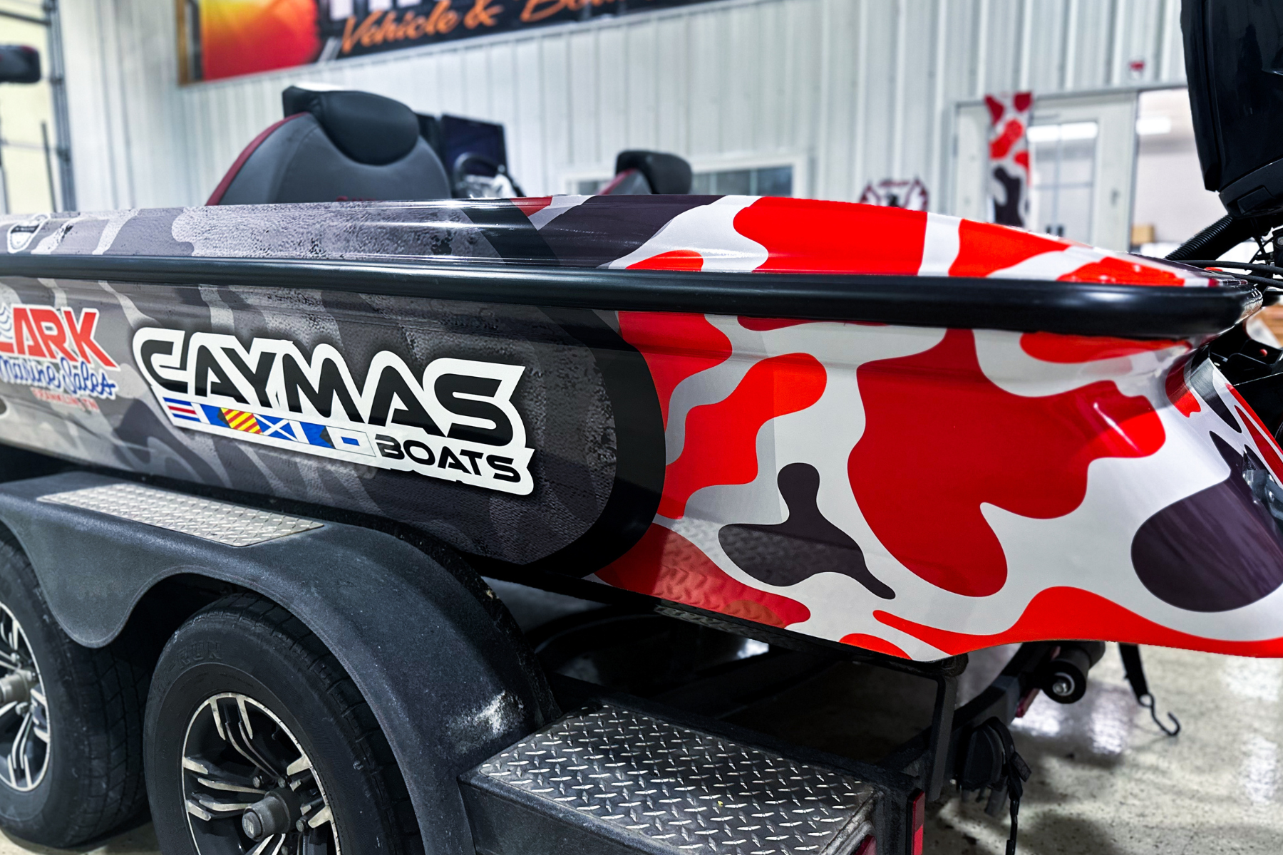 Red, black, and white camouflage-patterned boat on a trailer inside a garage with promotional banners on the wall.