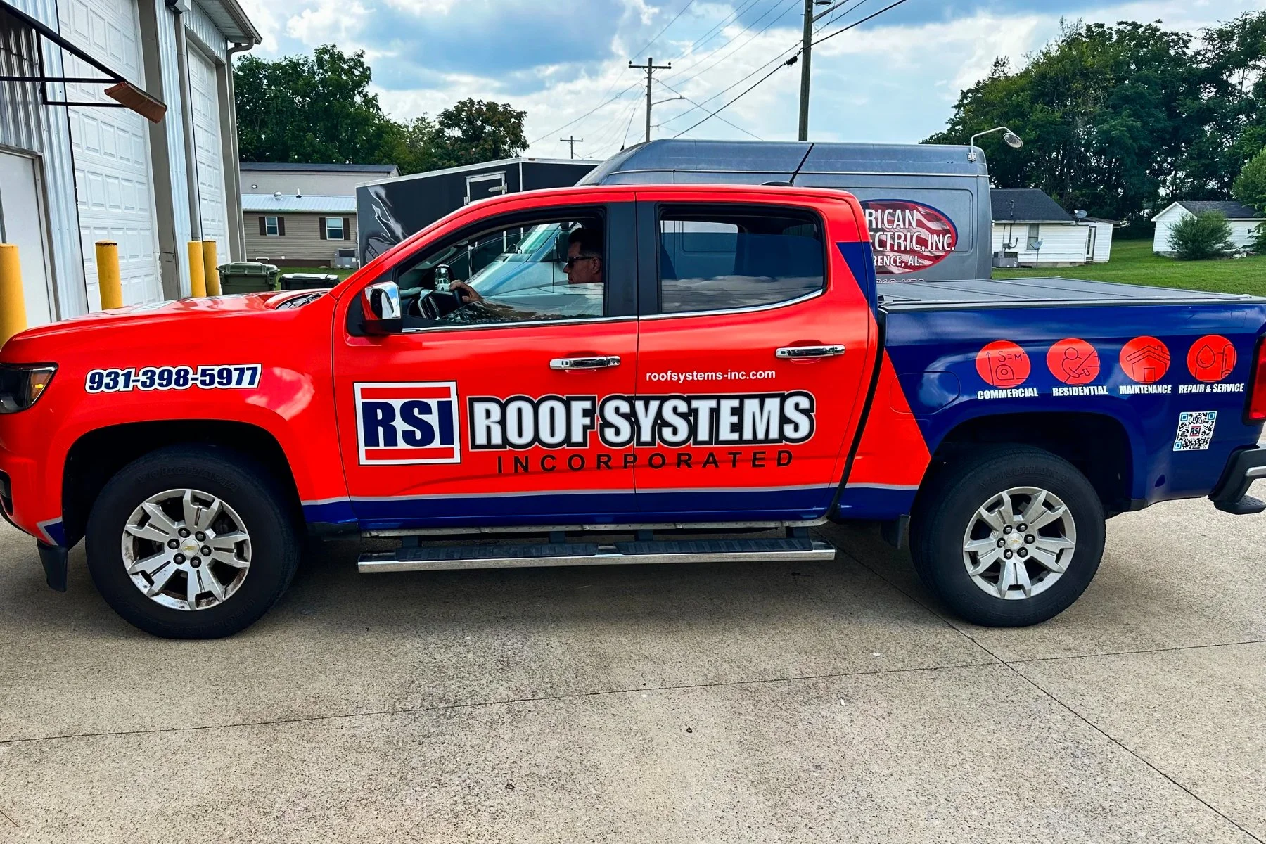 Red and blue pickup truck with the logo and branding of RSI Roof Systems on the side, parked outdoors near a building with a white wall and garage doors.
