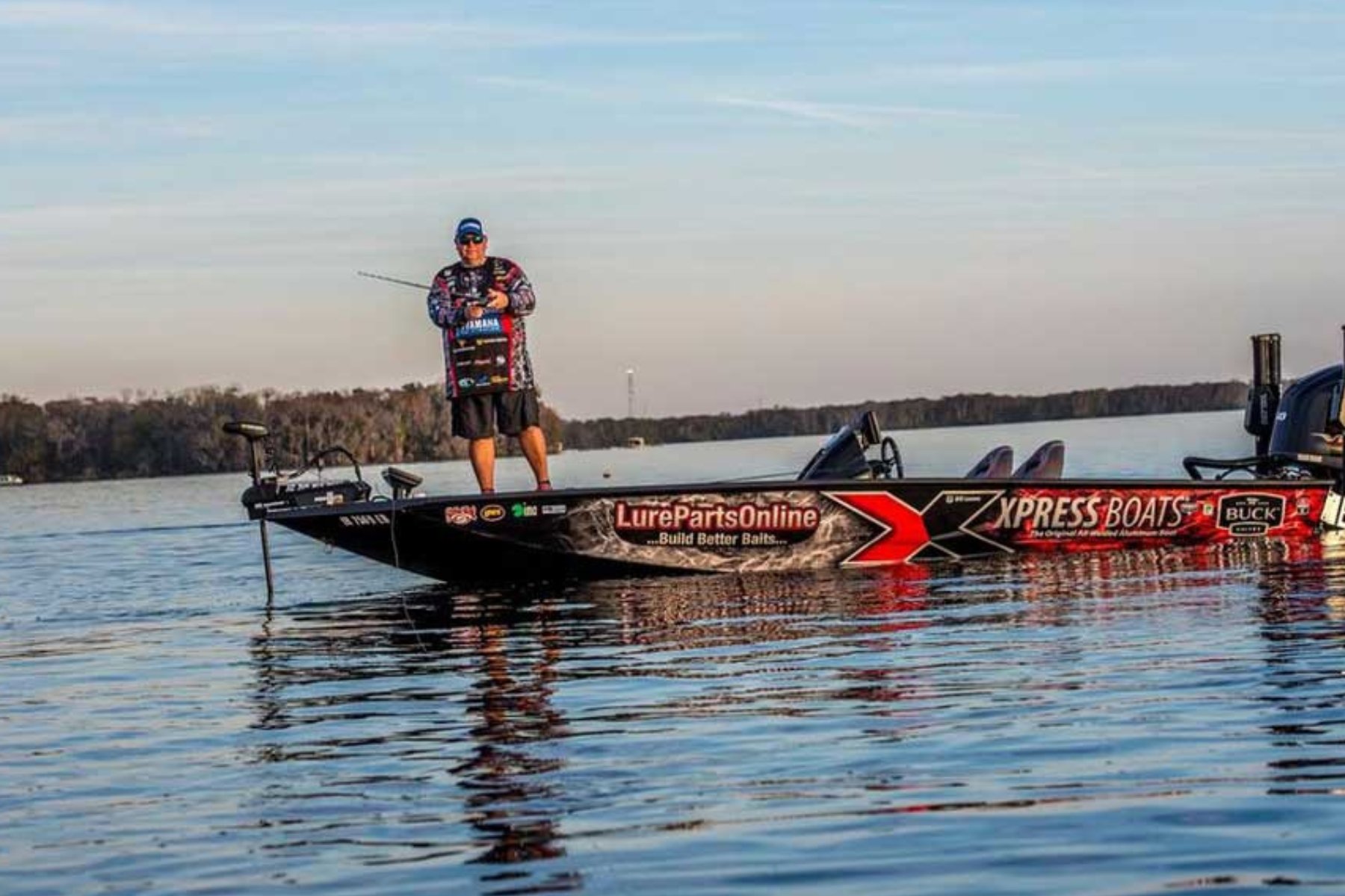 A man standing on a fishing boat on a lake, holding a fishing rod with a calm water and distant treeline in the background.