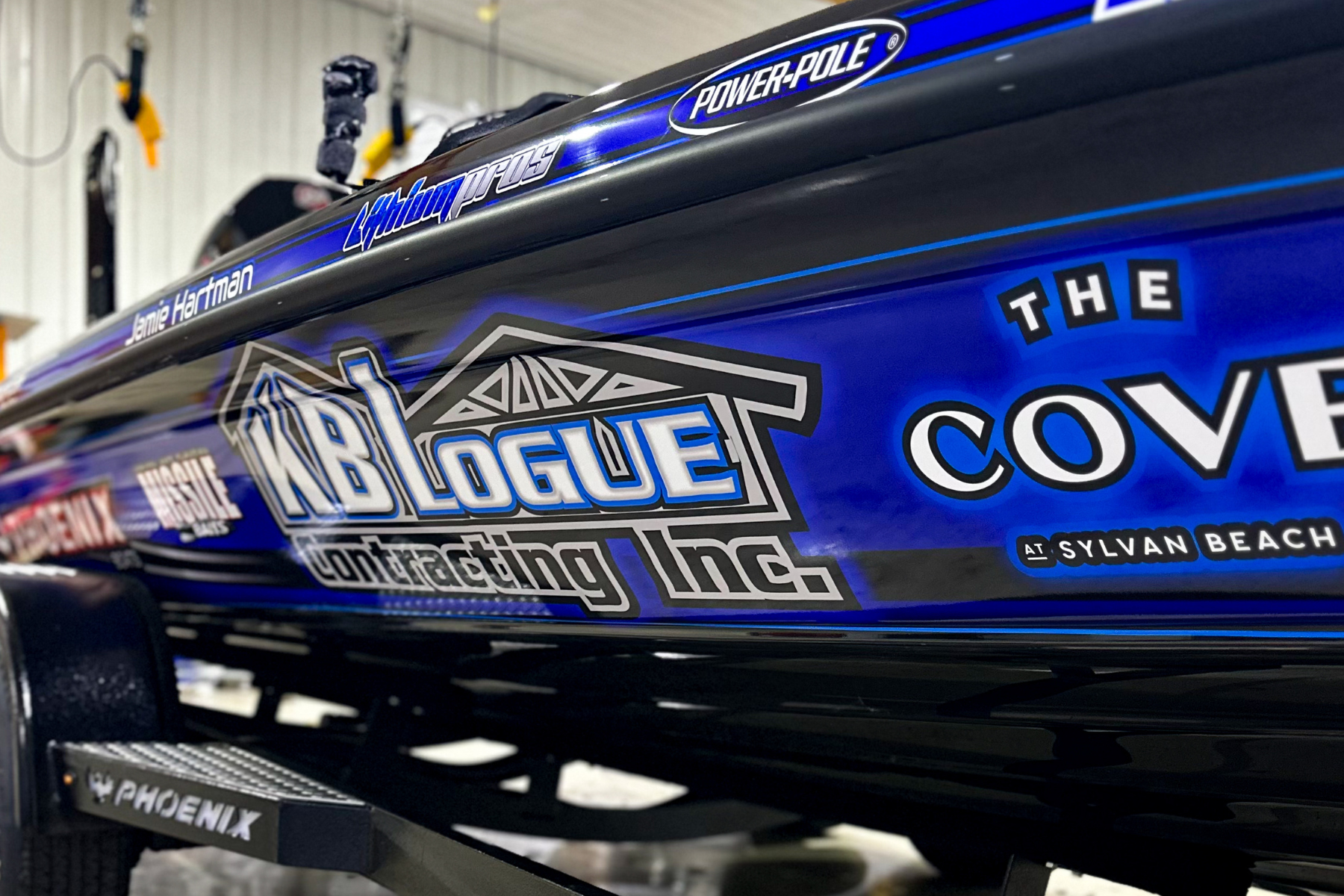 Close-up of a boat with blue and black paint, featuring logos including KB Logo Contracting Inc., The Cove at Sylvan Beach, and Power-Pole, in a garage or workshop setting.