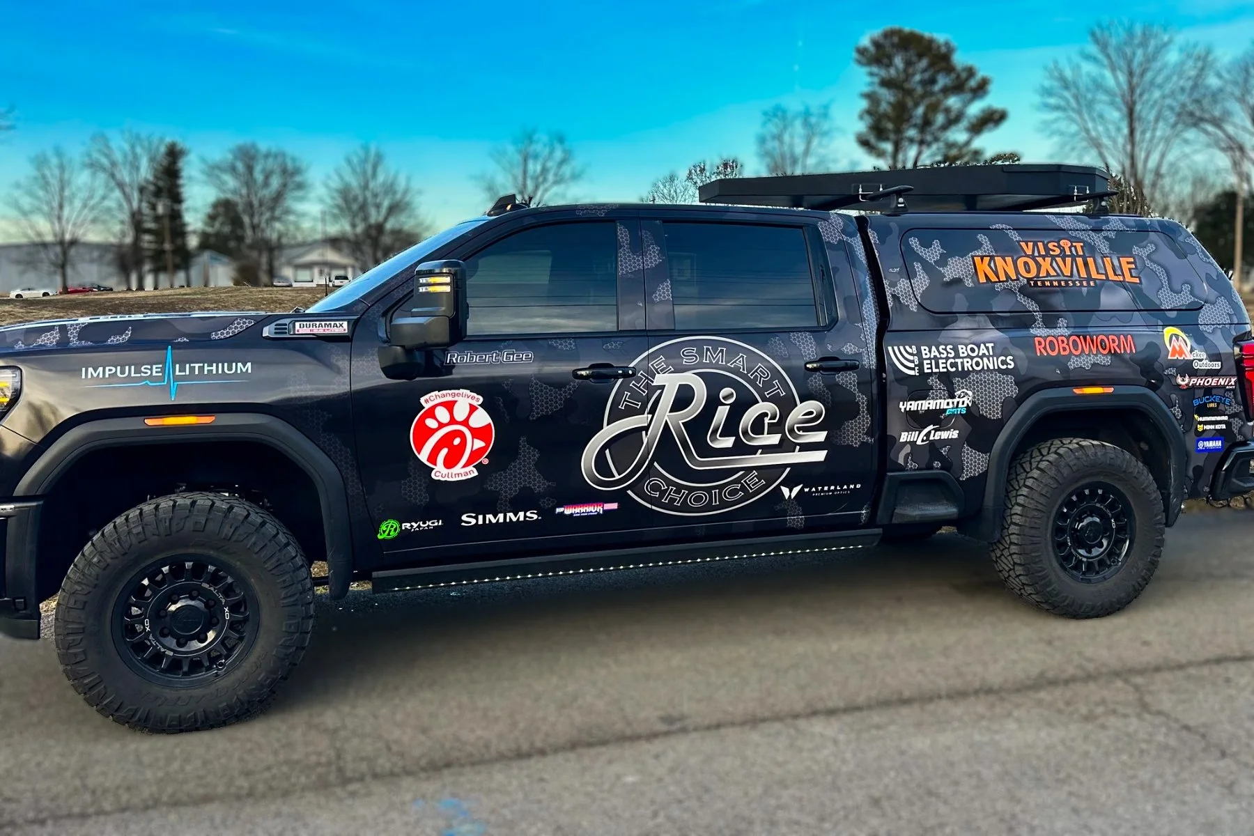 A black pickup truck with various sponsor logos and decals on its sides, parked outdoors on a paved surface with trees and a blue sky in the background.
