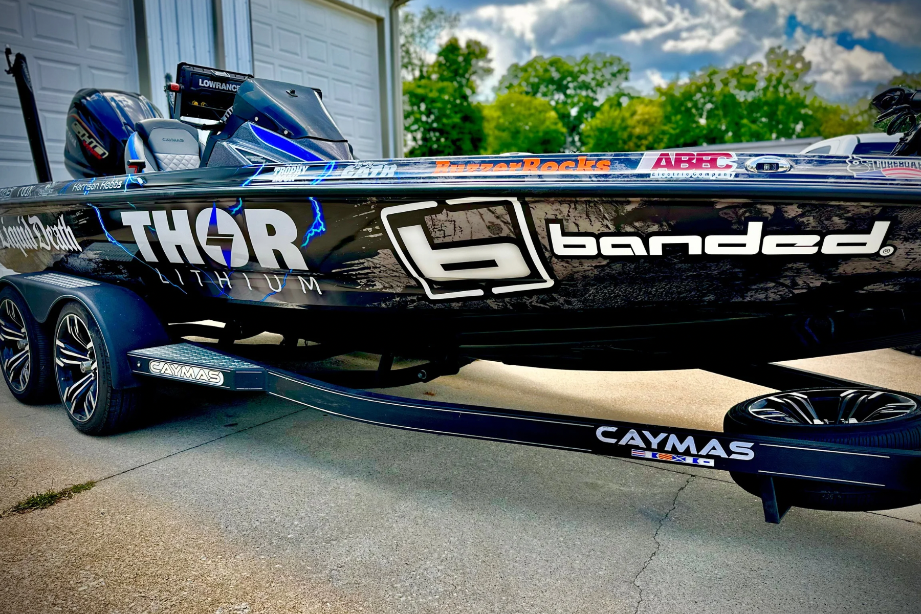 A black bass boat with various sponsor stickers, mounted on a dual-axle trailer on a concrete driveway, with a garage door and green trees in the background.