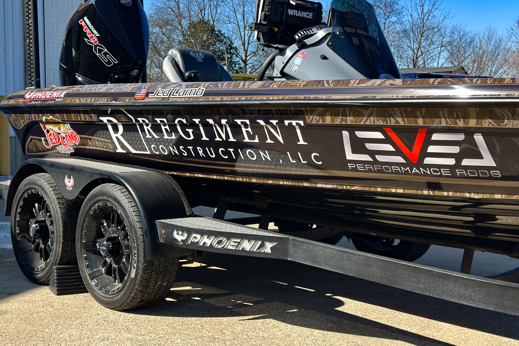 A bass boat with various sponsor decals parked on a trailer, including brand names like Phoenix and Mercury, with camouflage and black colors, under a clear blue sky.