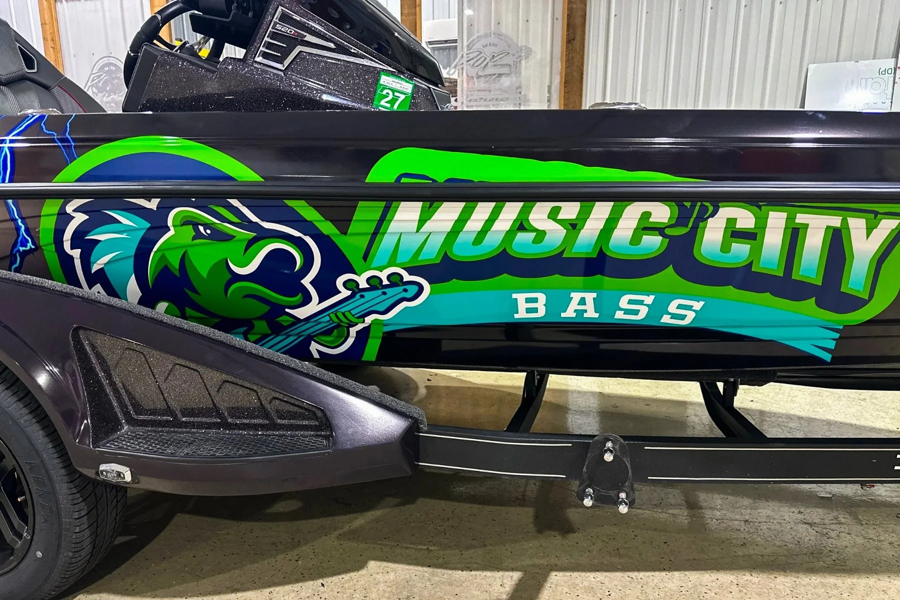 Colorful bass boat with a graphic of a green and blue lion holding a bass guitar, along with the text 'Music City Bass' in bright colors, parked indoors.