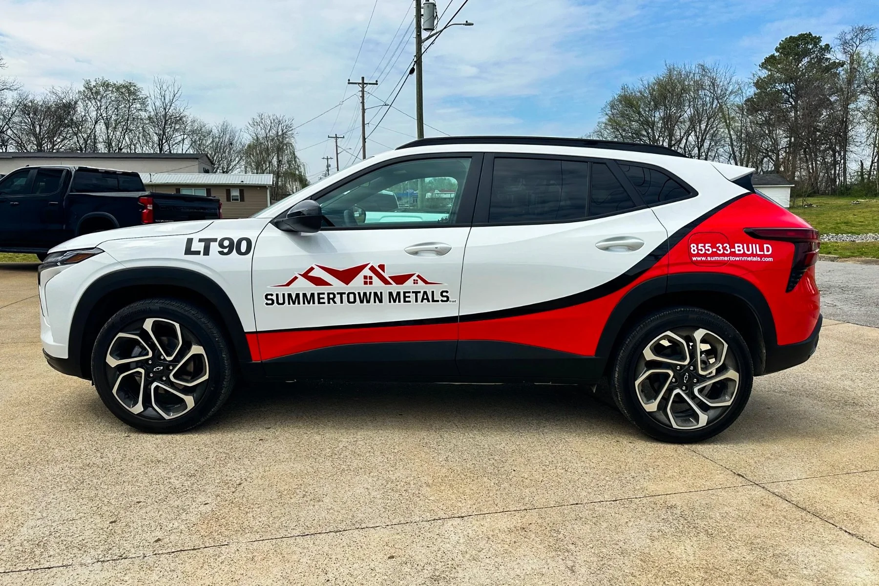 White SUV with red and black graphics, promoting Summertown Metals, parked on a paved driveway with a blue sky and trees in the background.