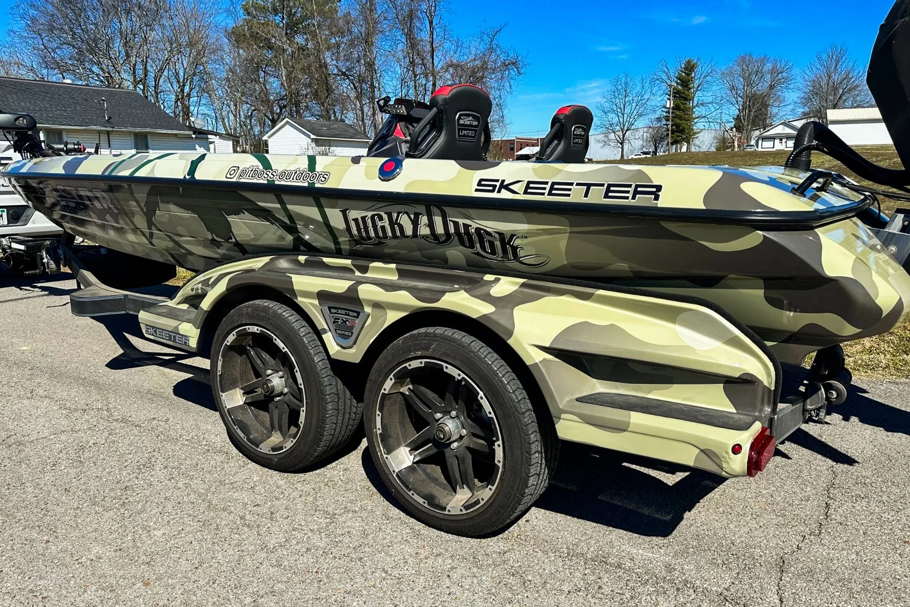 A camouflage-patterned Skeeter fishing boat on a matching dual-axle trailer, parked on a paved surface with houses and leafless trees in the background, under a clear blue sky.