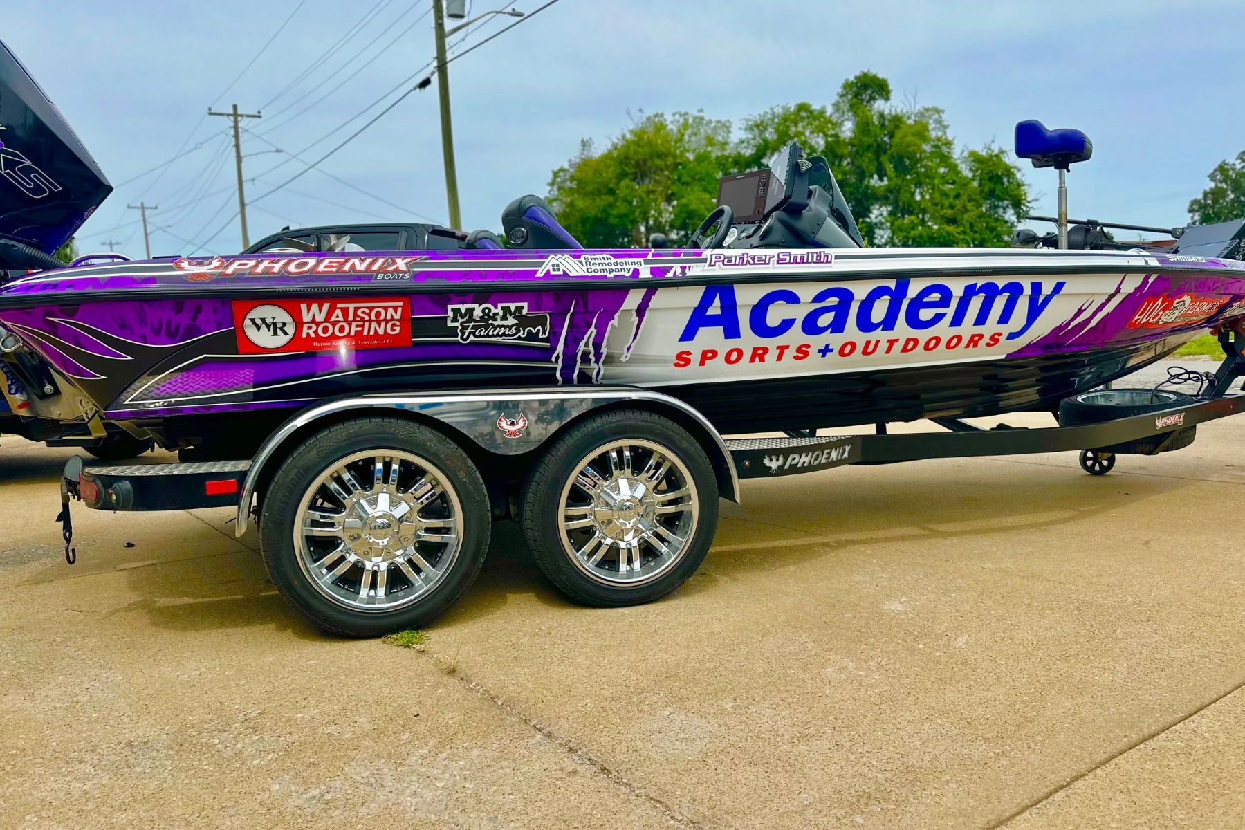 A purple and white boat trailer with a fishing boat on it, featuring various sponsor logos, parked on a concrete surface with power lines and trees in the background.