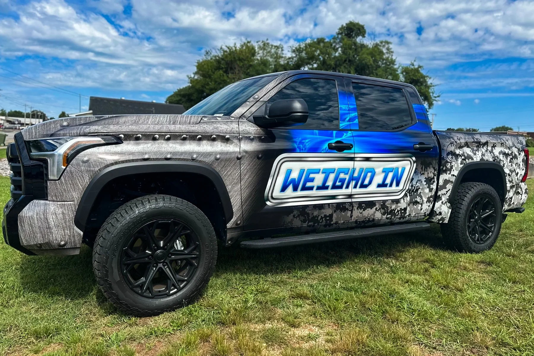 A pickup truck with a camouflage and graffiti-style design, featuring a large sign on the door that says 'WEIGH IN' in bold blue letters. The truck is parked on grass under a partly cloudy sky.